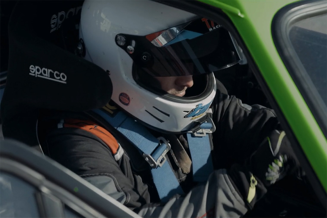 Race car driver wearing a white helmet with a blue harness and black racing gloves gripping the steering wheel inside a green sports car.