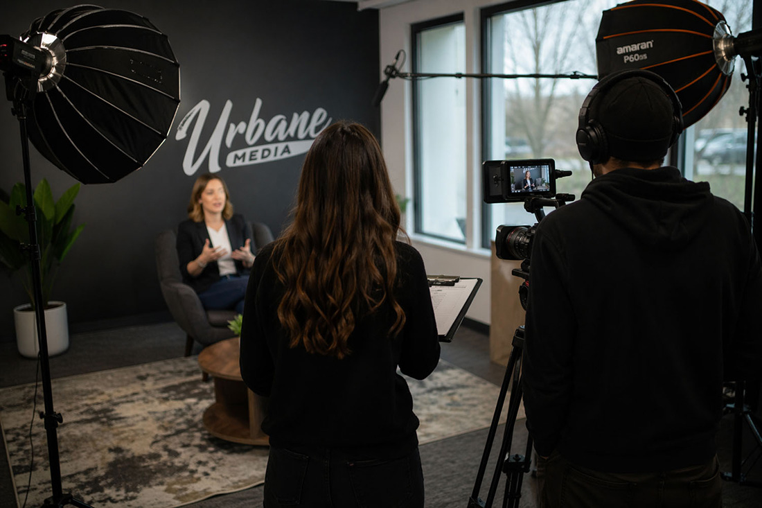 A woman is being recorded on video in a studio with 'Urbane Media' on the wall behind her as two crew members operate a camera and take notes under professional lighting.
