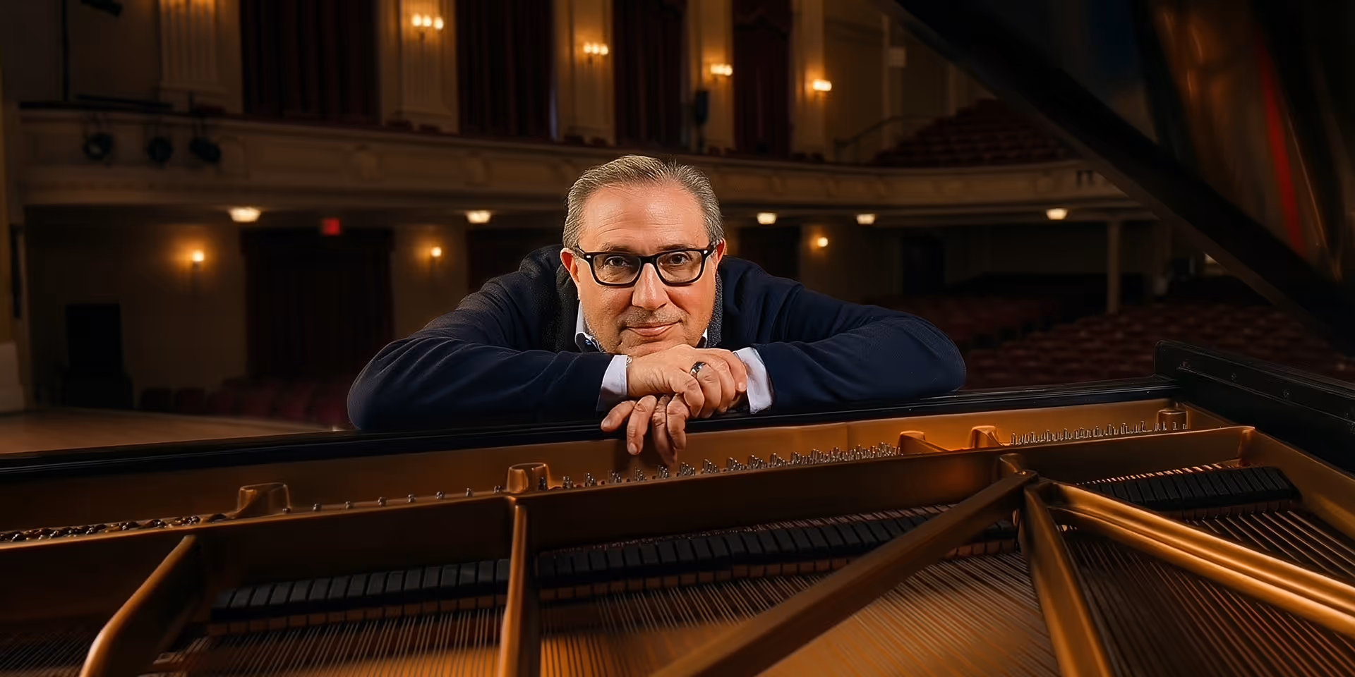 Man wearing glasses leaning on a grand piano inside a dimly lit concert hall.