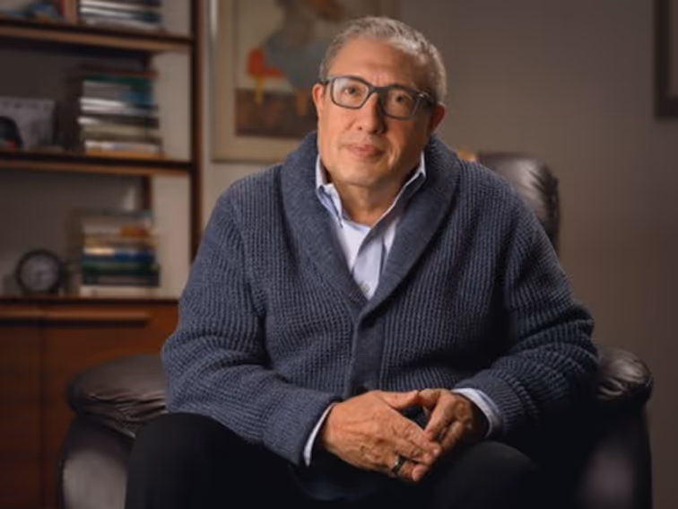 Middle-aged man wearing glasses and a blue knit sweater sitting in a leather chair with hands clasped, with a bookshelf and framed picture in the background.