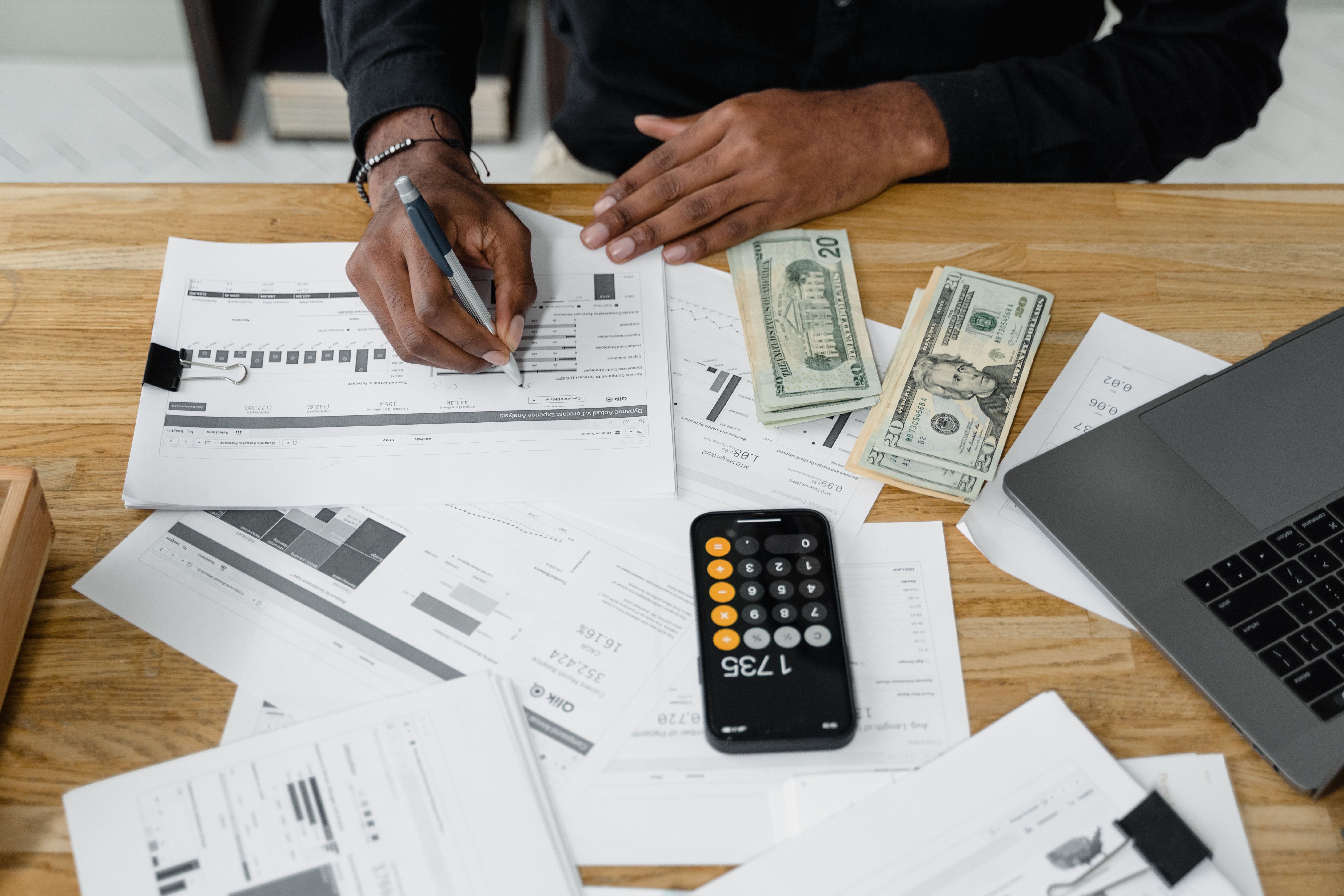 Person reviewing financial documents and charts at a desk with cash, a calculator, and a laptop nearby.