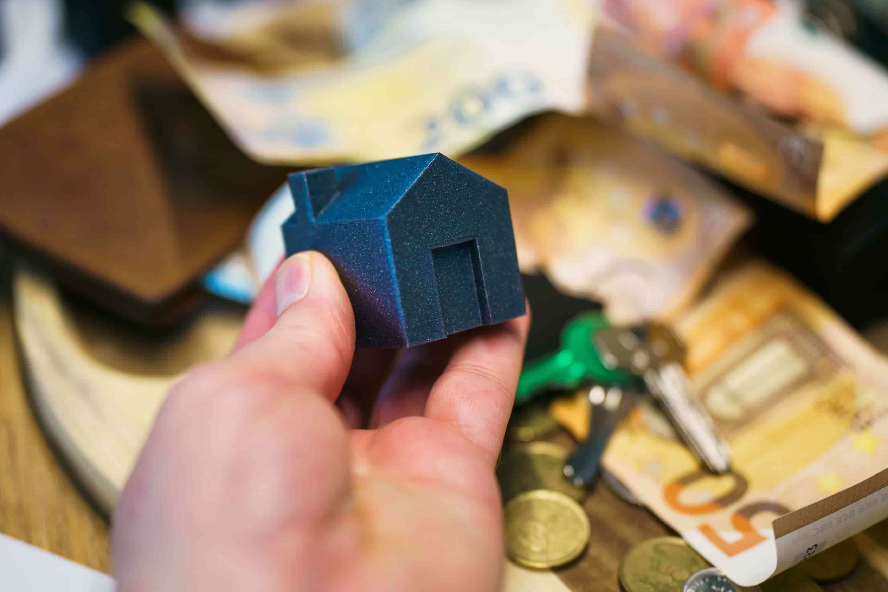 A hand holds a small dark blue miniature house figurine, with euro banknotes, coins, keys, and more tiny house models scattered on a wooden surface in the background.