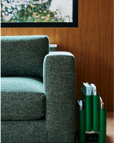 Close-up of a green upholstered sofa armrest beside a green magazine holder containing books, set against a wood-paneled wall and window.