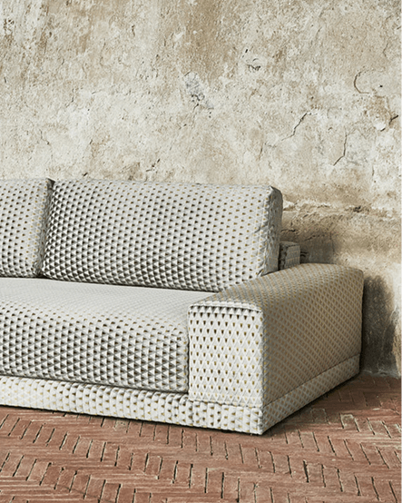 Modern light-gray textured sofa placed against a rough, weathered concrete wall on a brick floor.