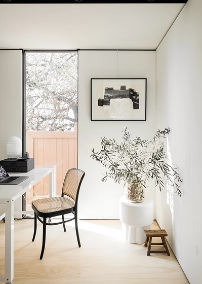 Minimalist room with a black-framed window, a white desk with a laptop, a woven chair, a framed abstract artwork on the wall, and a large vase with leafy branches on a white pedestal next to a small wooden stool.
