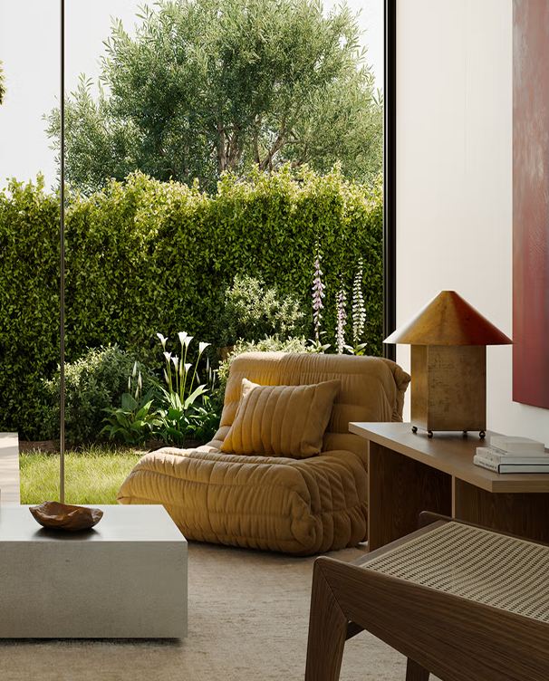 Cozy modern living room corner with a plush brown lounge chair, matching pillow, wooden side table with lamp, and large window showing green garden outside.