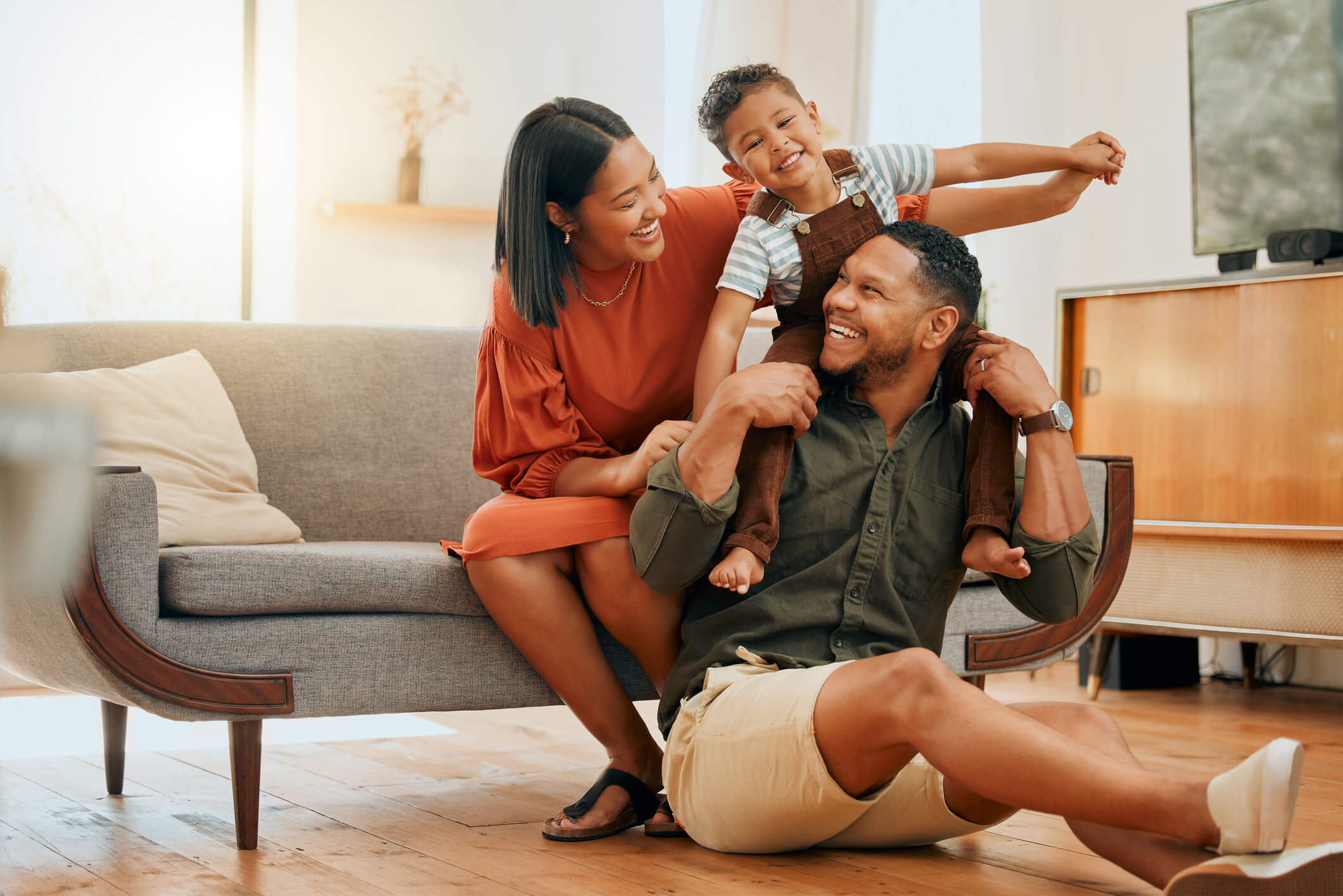 Smiling family of three playing together in a cozy living room with the child sitting on the father's shoulders.