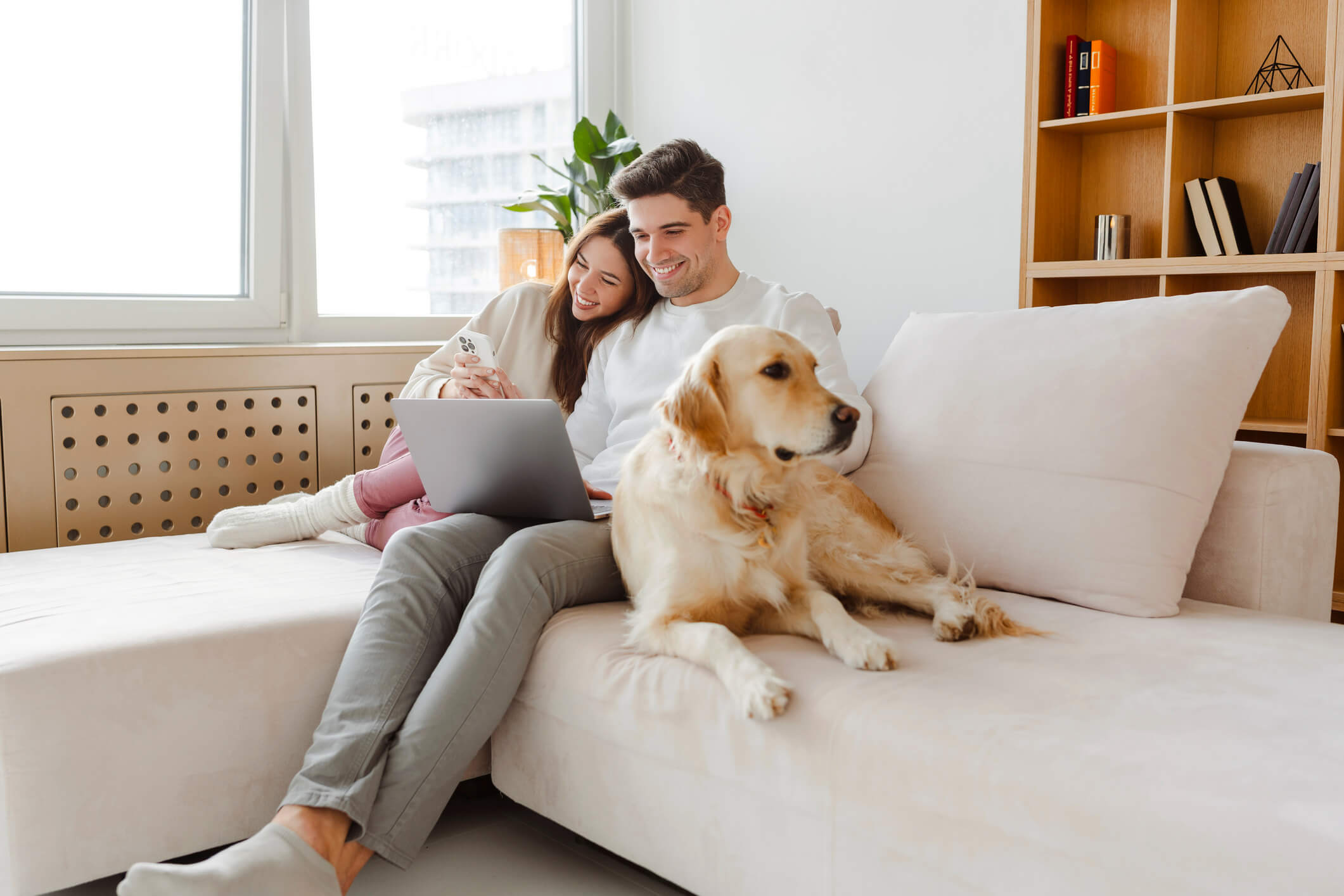 Smiling couple sitting on a beige couch with a laptop and smartphone, a golden retriever lying beside them.