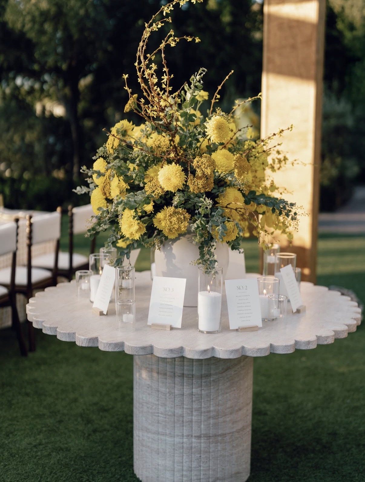 Round white table with a large yellow floral arrangement centerpiece, surrounded by small candles and table number cards.