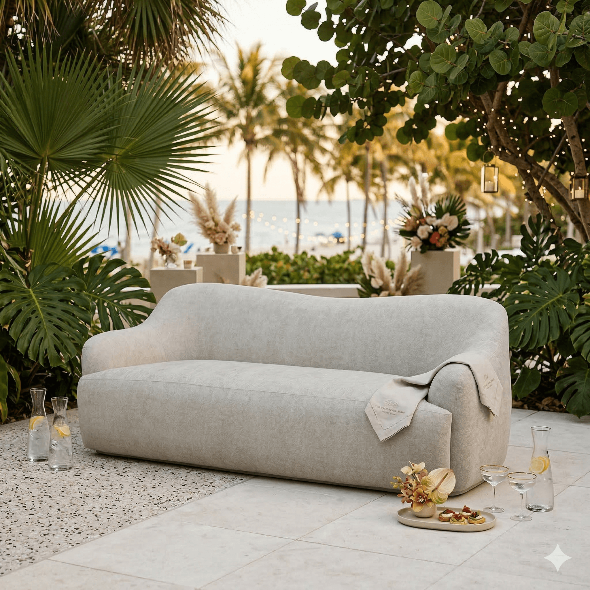 Modern beige sofa on a patio with tropical plants, decorative flowers, and a tray with drinks and appetizers, ocean and palm trees in the background.