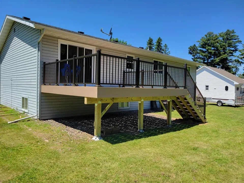 Elevated wooden deck with black metal railing and stairs attached to a gray house under a clear blue sky.