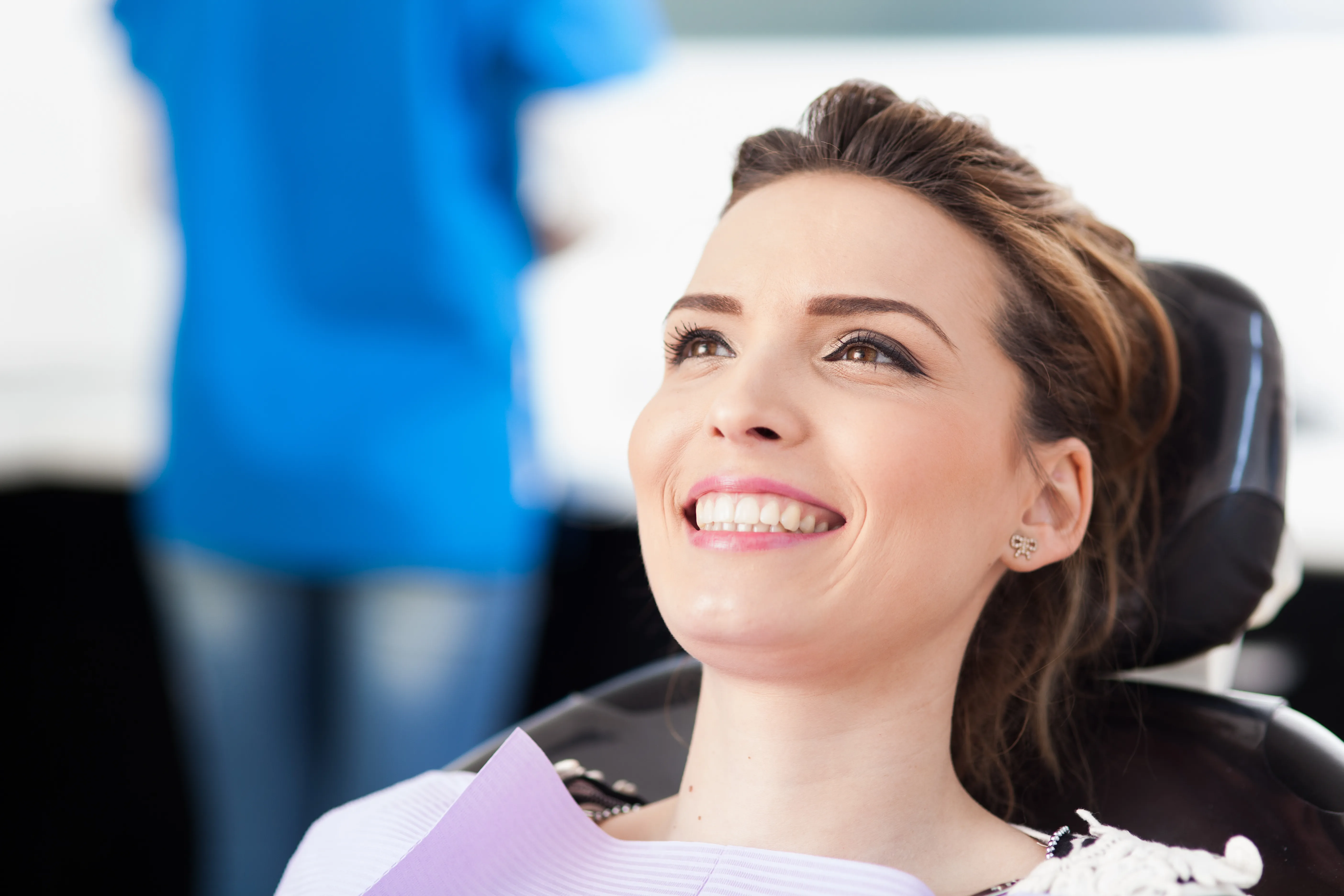 Young woman reclined in a dental chair smiling and looking up, with a clinician blurred behind her