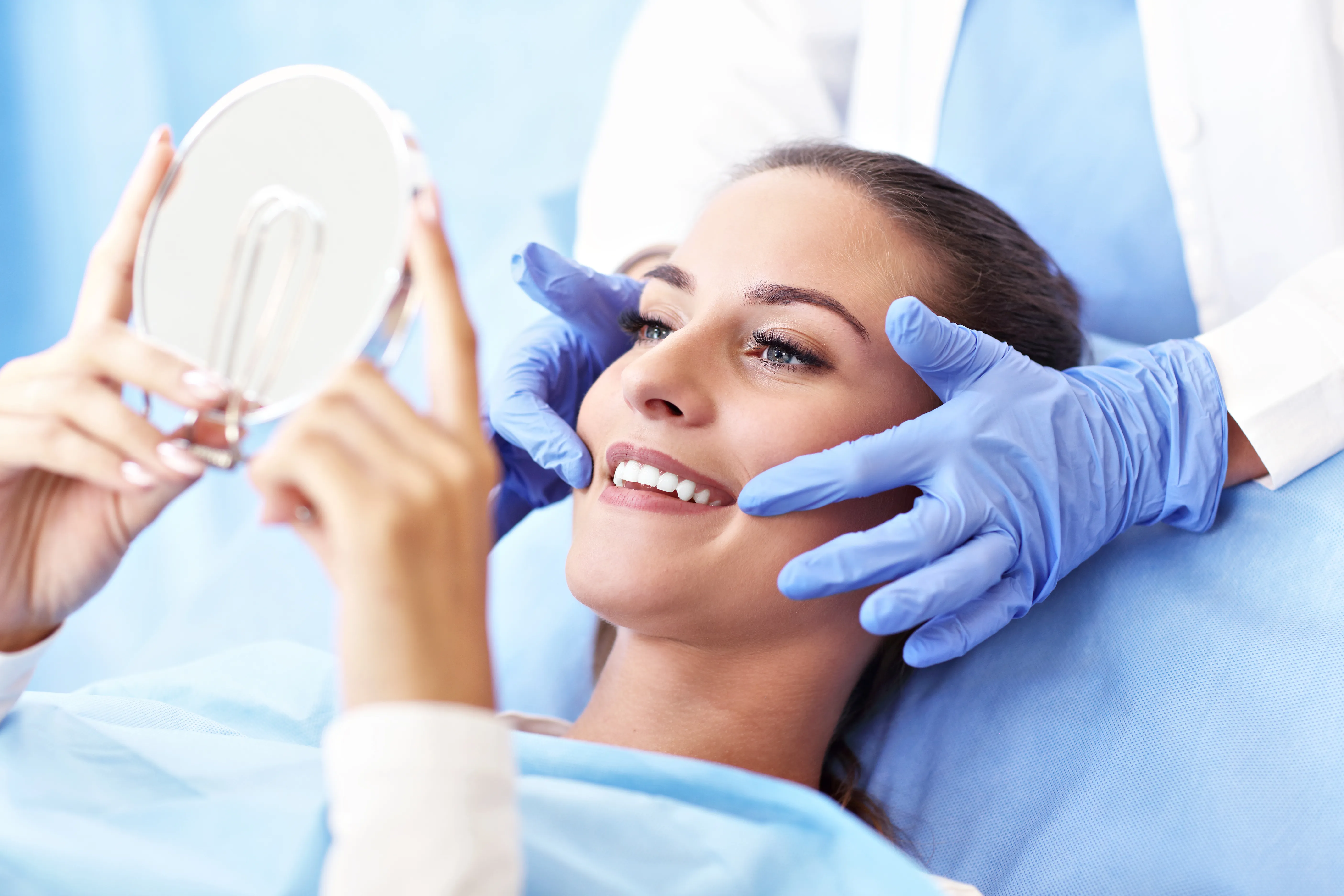 Woman in a dental chair smiling as she looks into a hand mirror held by a gloved dentist