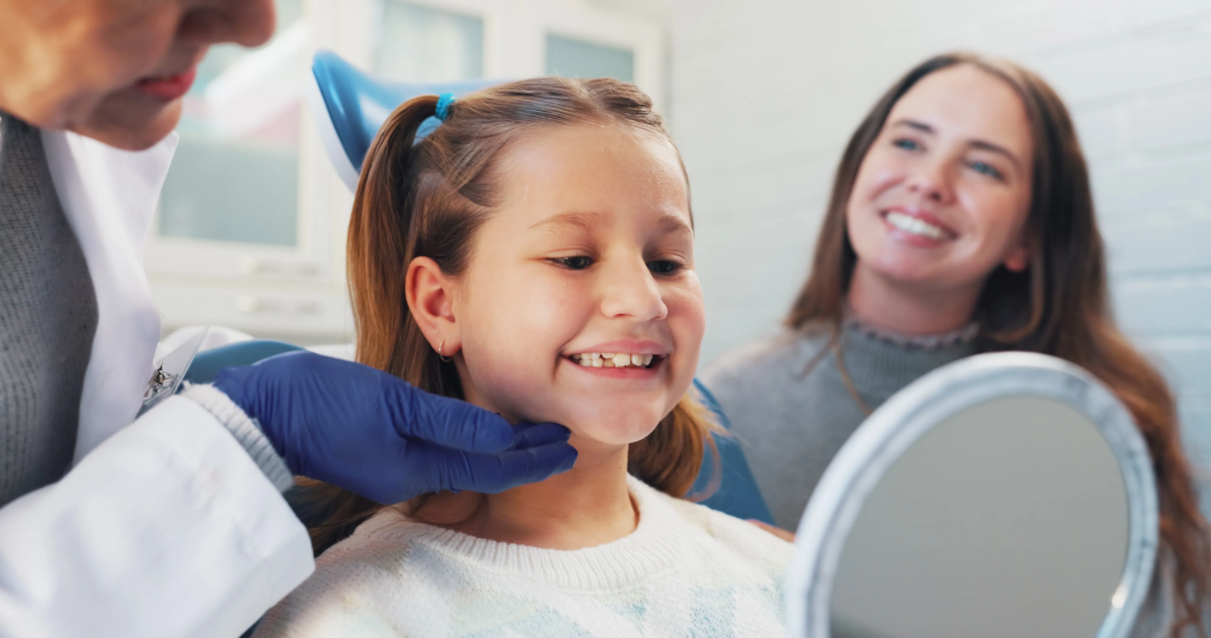 Smiling girl admiring her teeth in a hand mirror at the dentist with her mother nearby