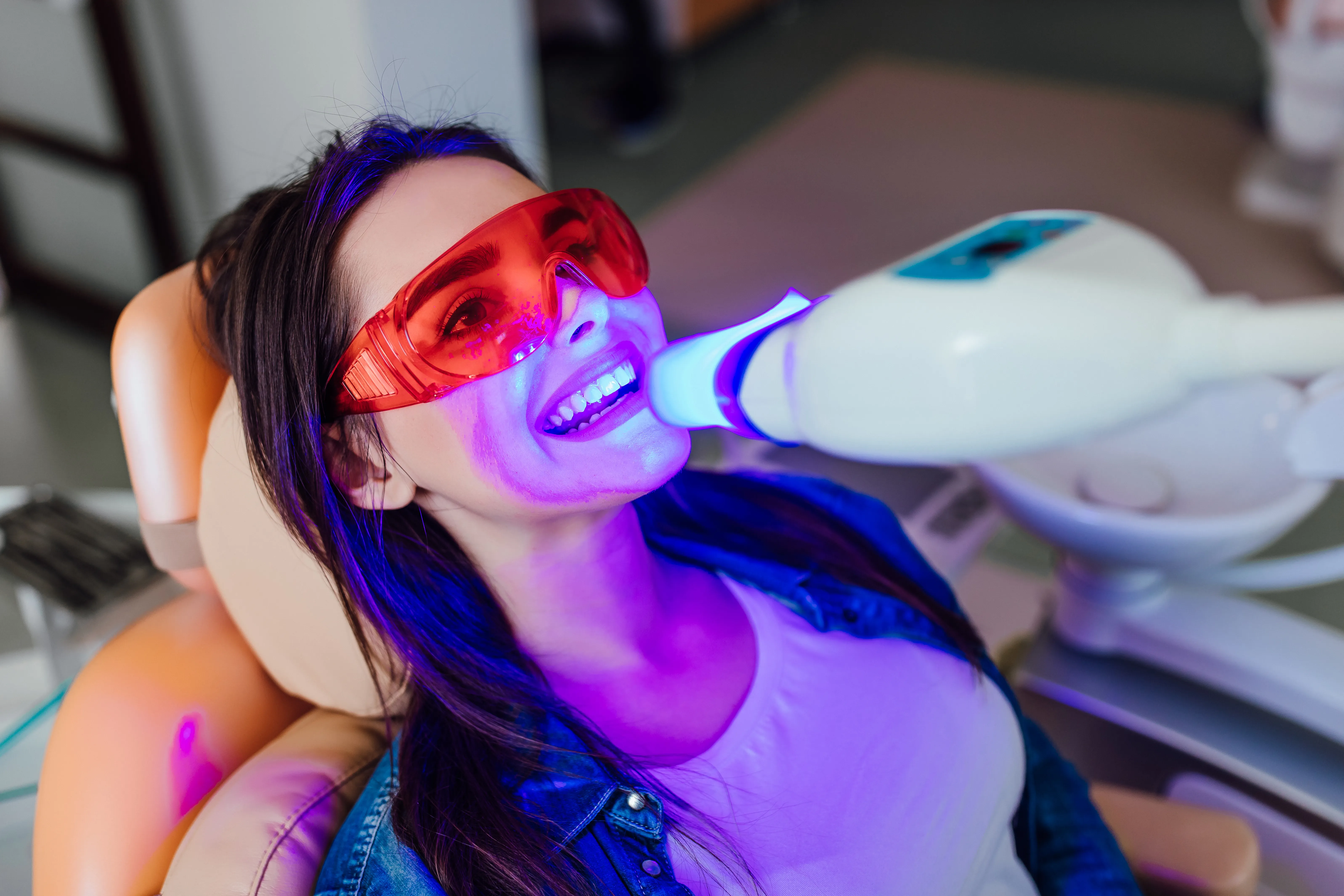 Woman in red protective glasses reclined in a chair during blue-LED teeth whitening with the lamp at her mouth
