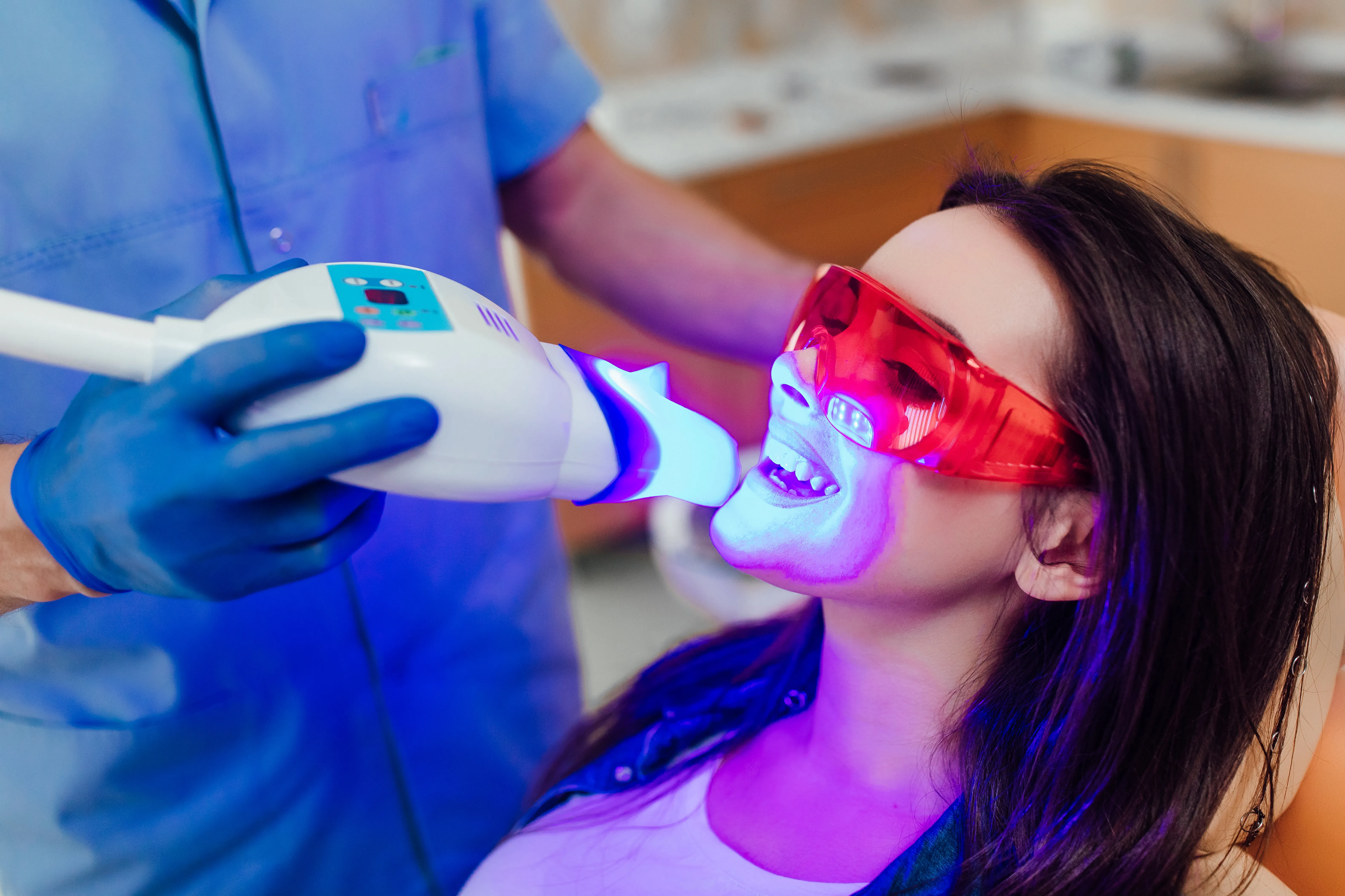 Woman in red protective eyewear receiving blue-LED teeth whitening as a gloved clinician holds the lamp