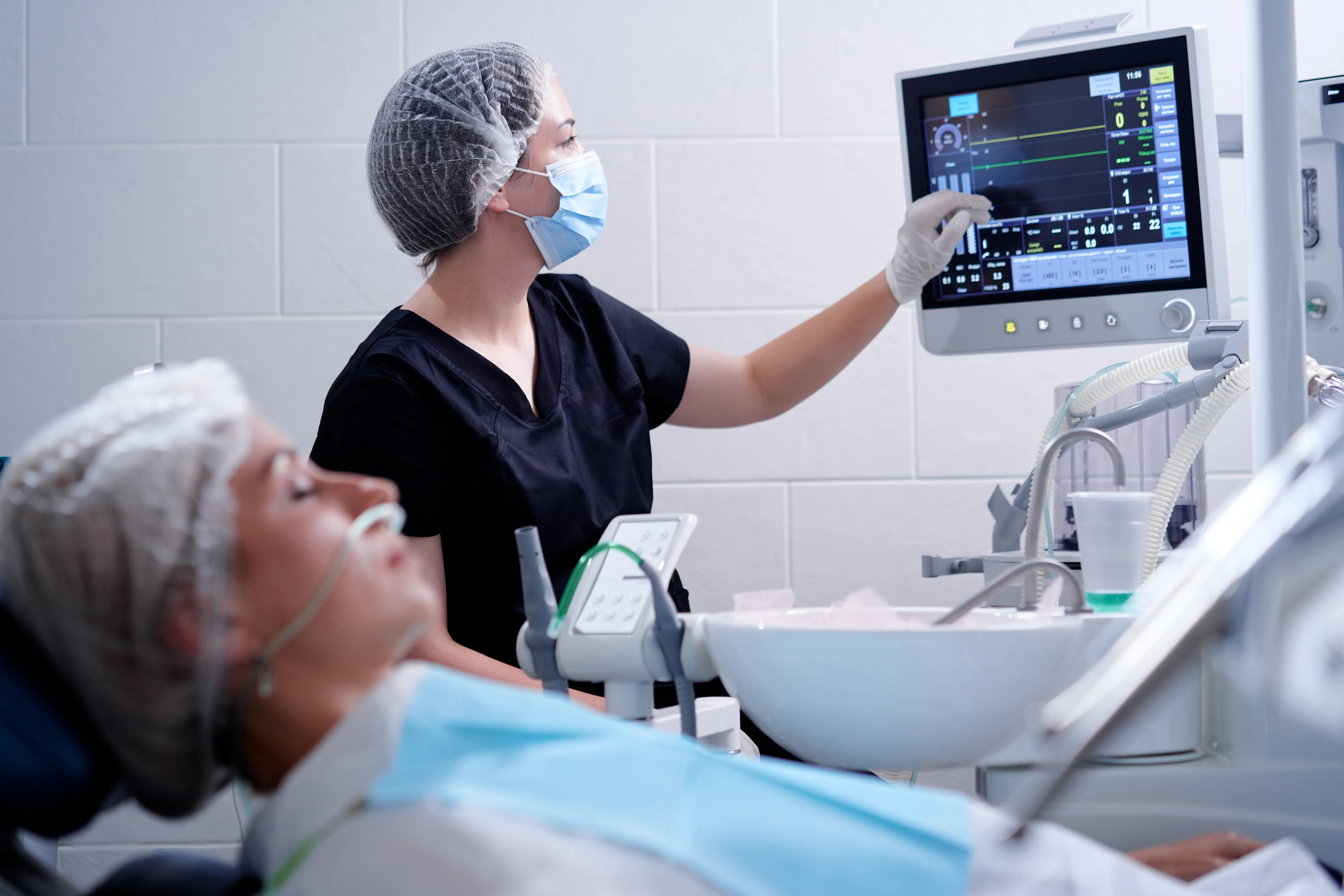 Sedation provider in scrubs adjusting a vital-signs monitor while a patient rests sedated with a nasal cannula