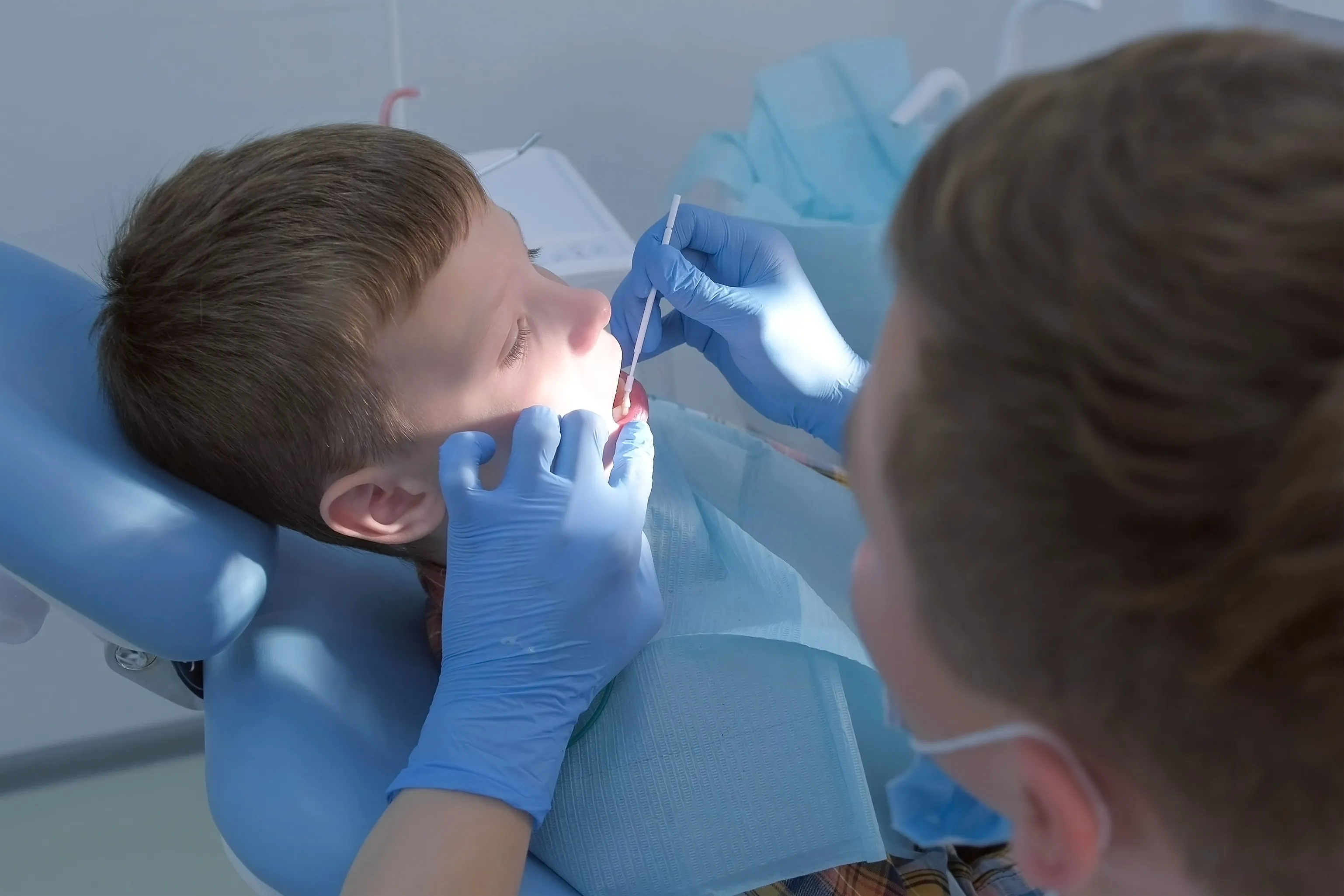 Young boy reclined in a dental chair while the dentist applies liquid to his teeth with a long-handled brush
