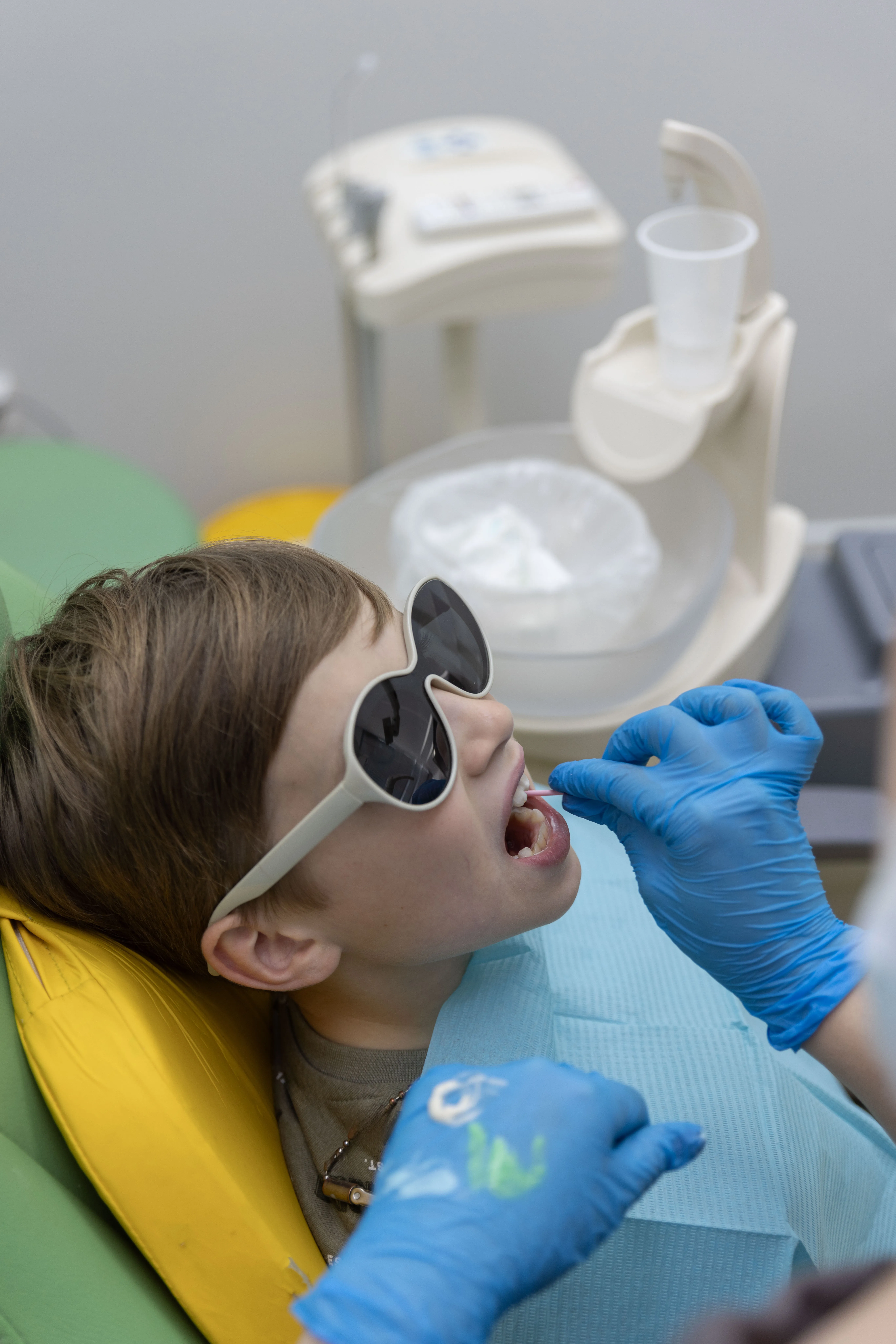 Child wearing protective glasses in a dental chair as a dentist applies liquid to their teeth with a small brush
