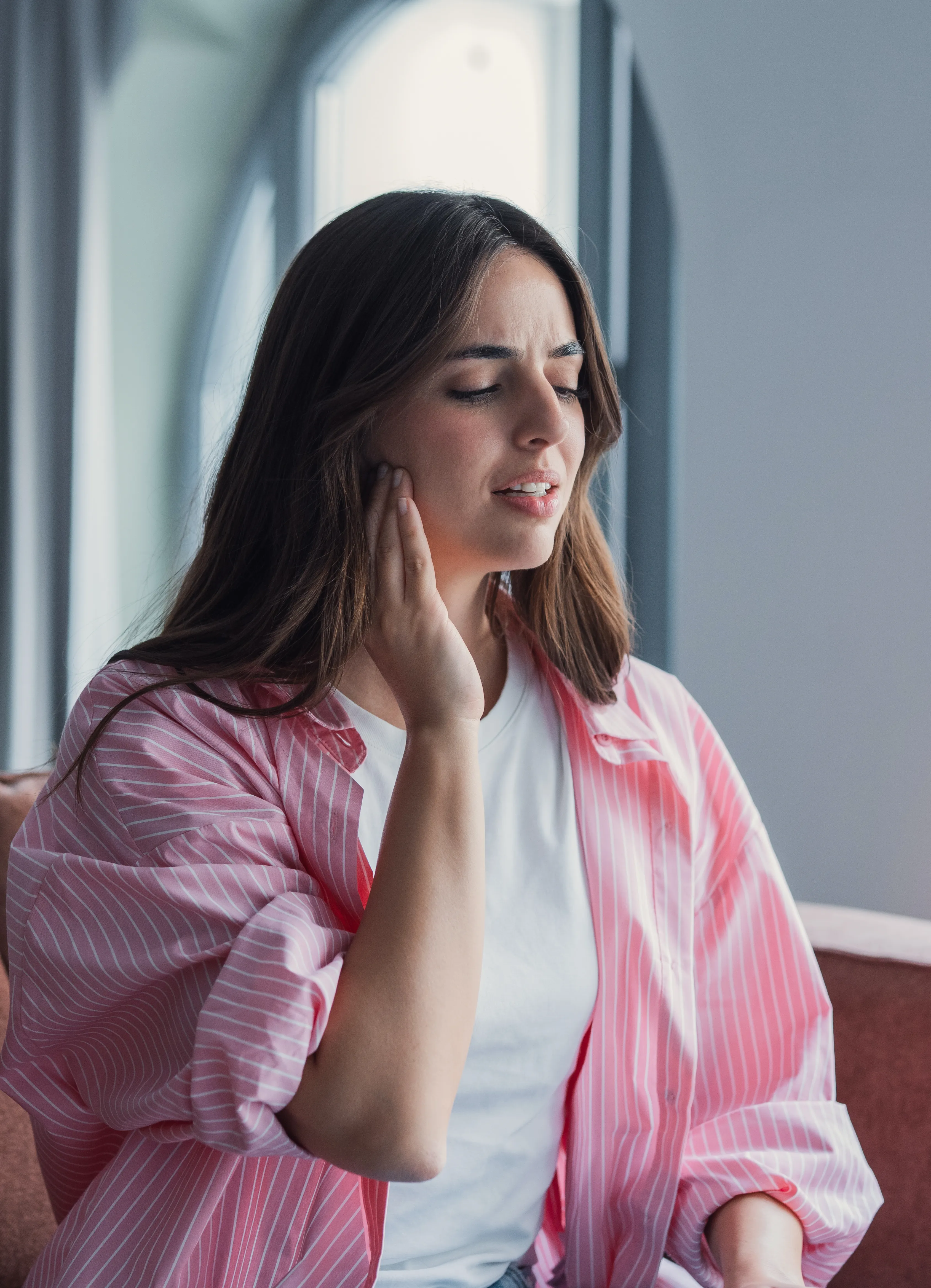 A young woman sits in a bright home interior, touching her jaw with a pained expression.