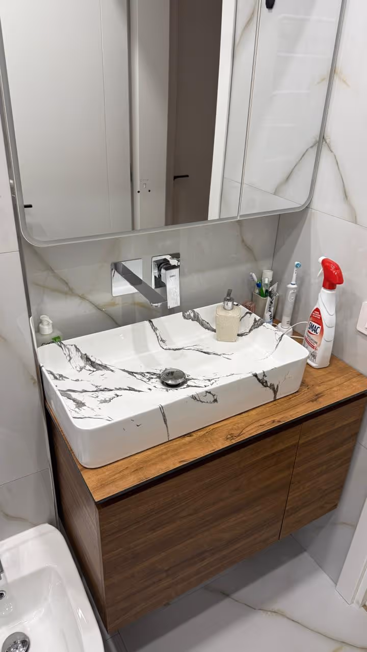 Lavabo me model guri mermeri mbi banakun prej druri me pasqyrë mbi të dhe produkte higjienike pranë qelqes dhe pasqyrës.