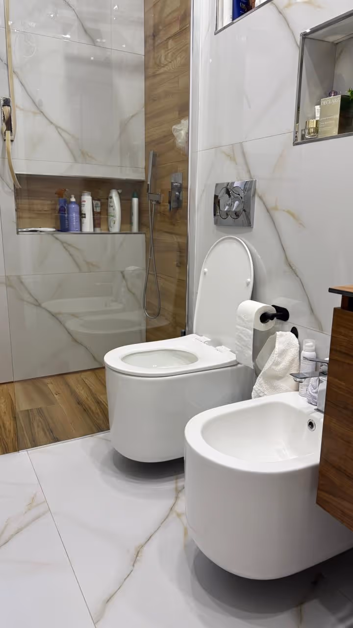 Banjo dhe bidé modern të bardhë në një banjë me pllaka mermeri dhe elementë druri.