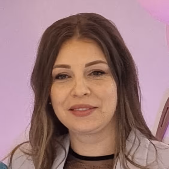 Grua me flokë kafe dhe të gjatë, duke buzëqeshur, përpara një sfondi rozë.