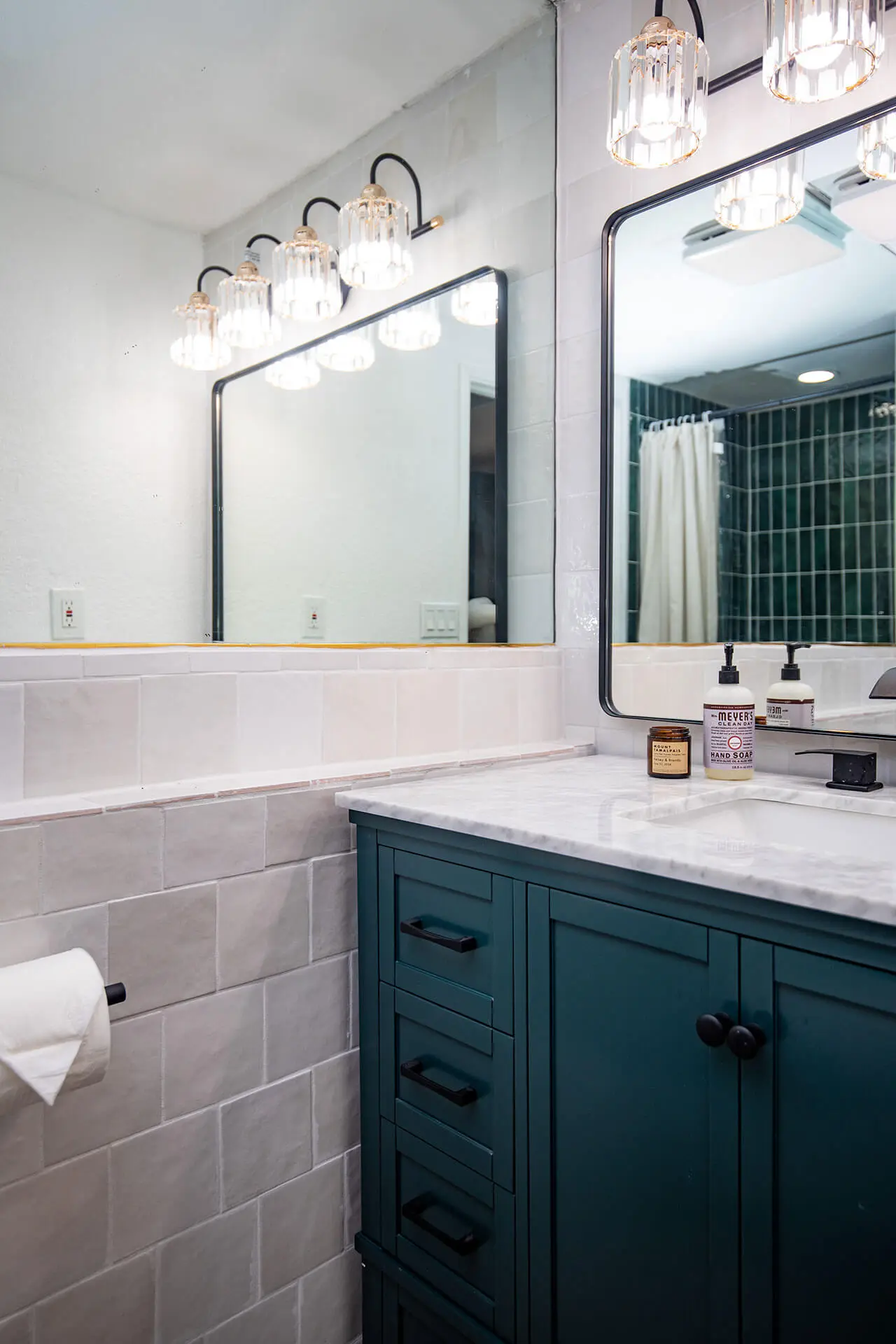 A renovated bathroom by Sunshine Collective Studio featuring a deep teal vanity with marble countertop, black hardware, and crystal globe sconce lighting above a black-framed mirror. White zellige subway tile lines the walls, with deep green tile visible in the shower reflection.