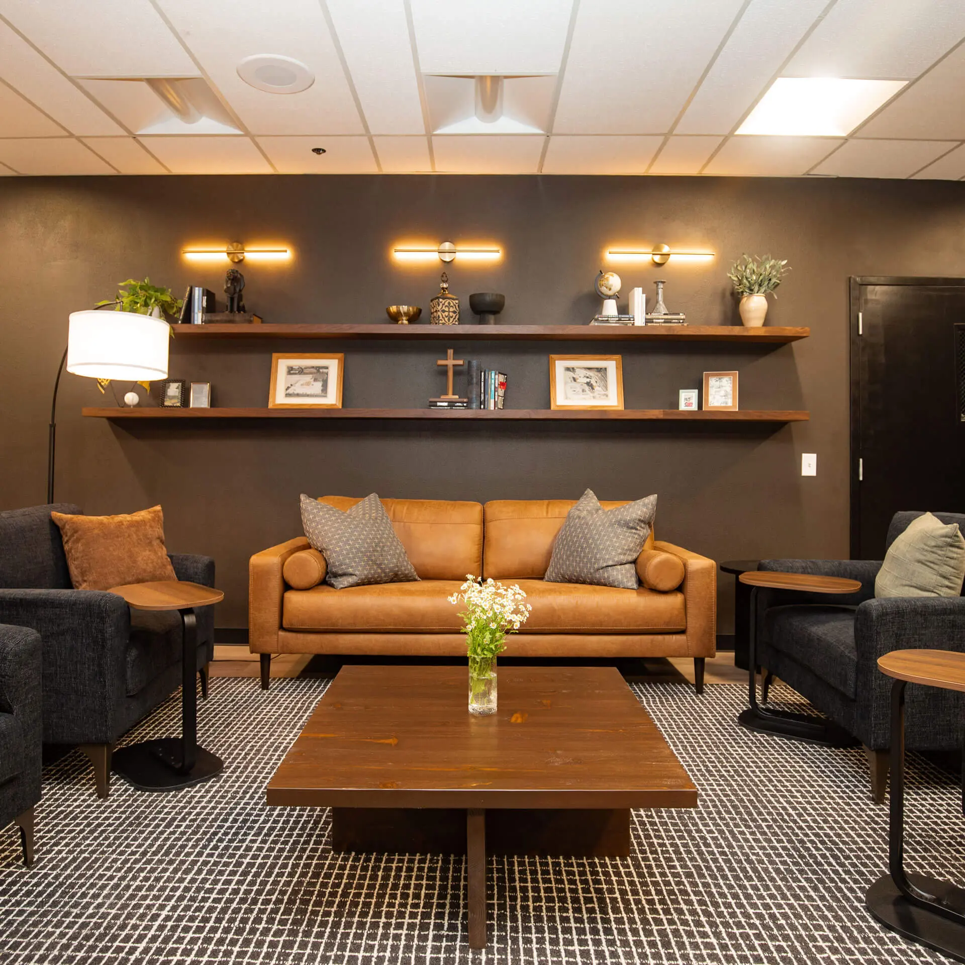 A commercial lounge space designed by Sunshine Collective Studio, featuring a cognac leather sofa flanked by dark charcoal armchairs, a solid walnut coffee table, and two tiers of floating walnut shelves displaying curated objects beneath warm brass sconce lighting against a deep charcoal accent wall.