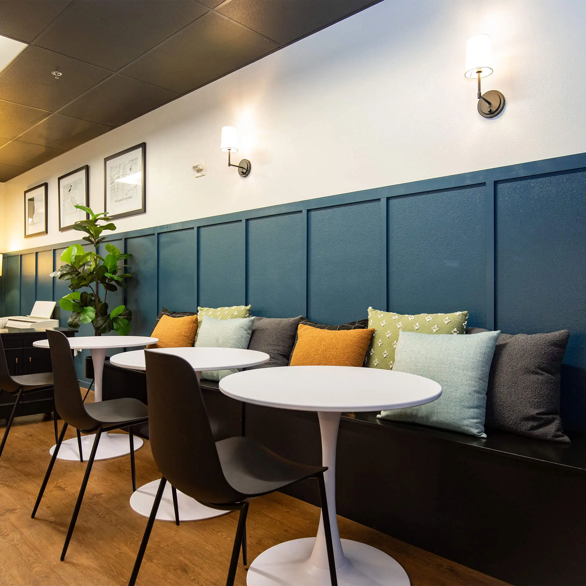 A refreshed commercial interior by Sunshine Collective Studio featuring a cushioned banquette bench along a teal board-and-batten accent wall, styled with layered throw pillows in amber, sage, and charcoal. White tulip tables with dark modern chairs line the seating area, alongside fiddle-leaf fig plants and framed map prints on warm wood flooring.