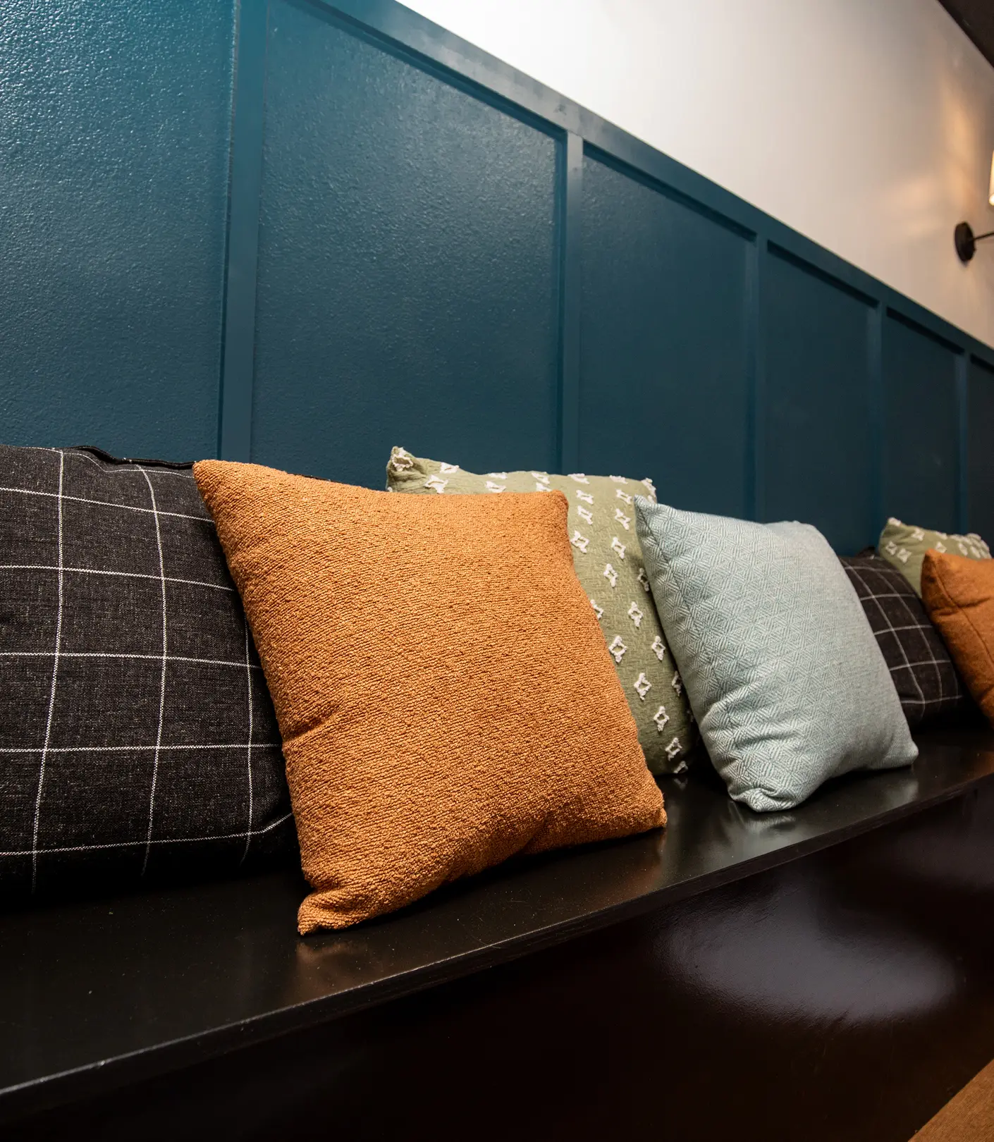 Banquette detail by Sunshine Collective Studio showing layered throw pillows in amber boucle, sage embroidered, light blue, and charcoal plaid against a teal board-and-batten wall.