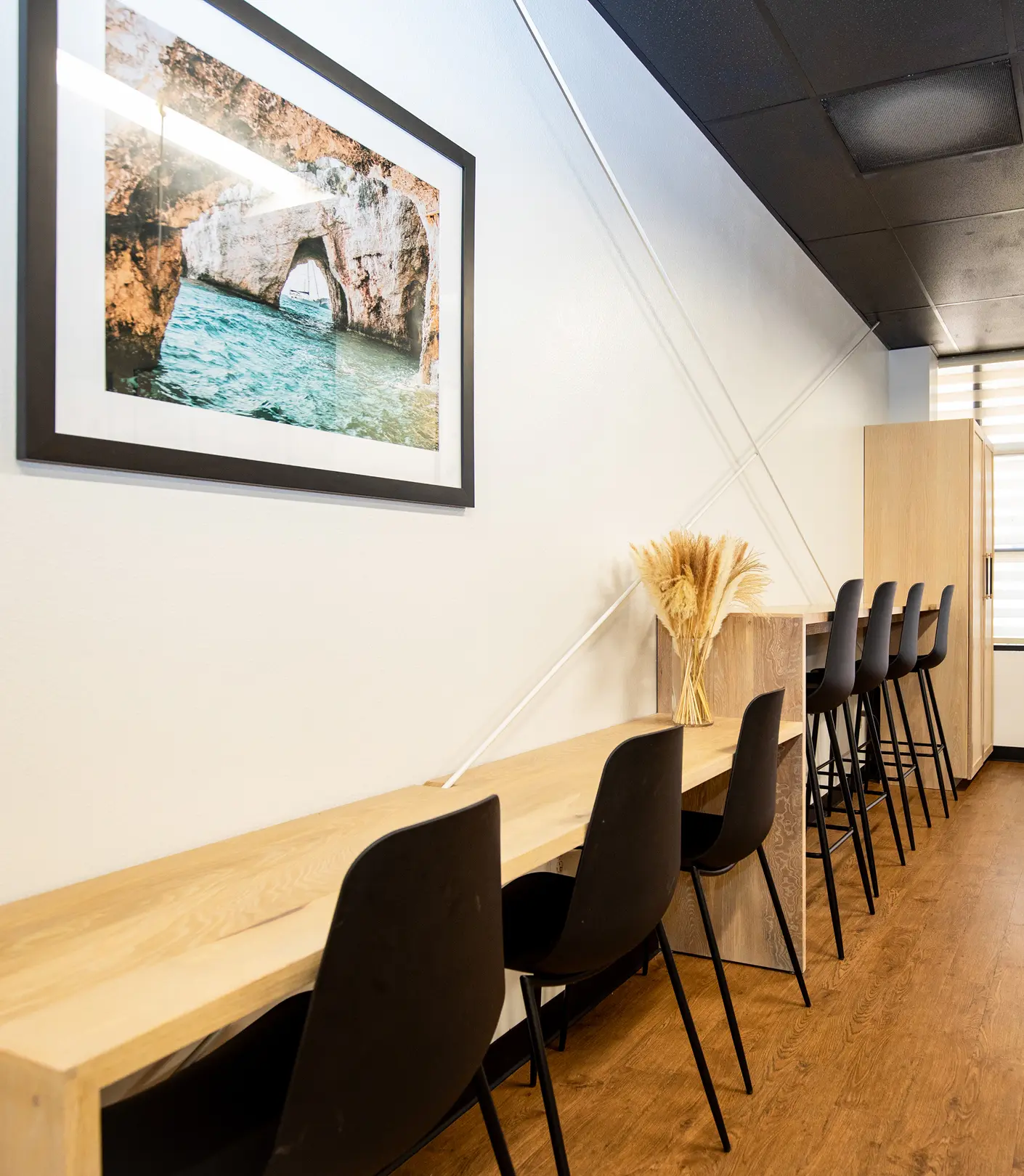 Workspace by Sunshine Collective Studio featuring a long oak counter, black bar stools, dried pampas grass, and a large framed coastal cave photograph on warm wood flooring.
