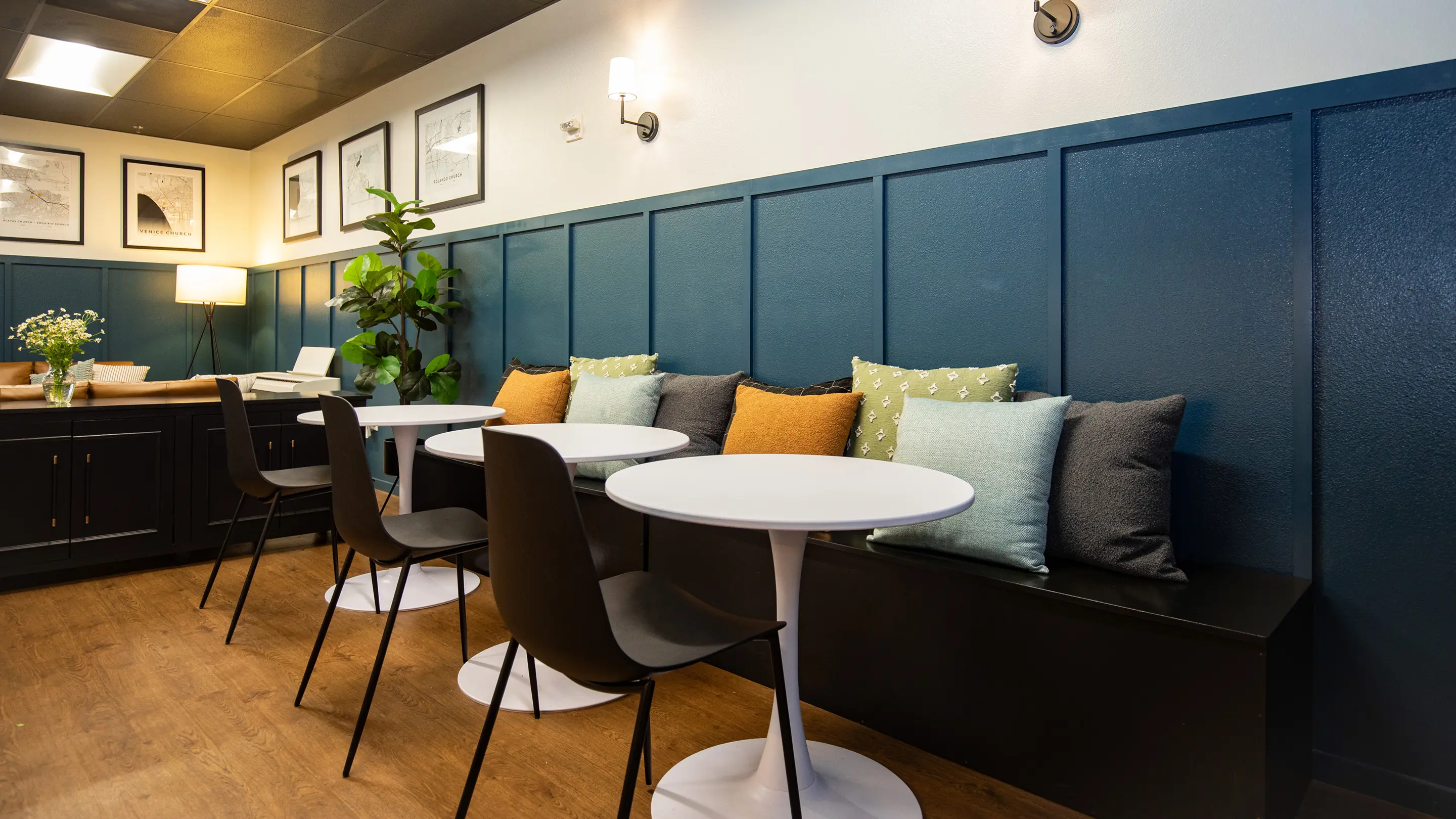 Eastlake lounge by Sunshine Collective Studio with a teal board-and-batten banquette styled with amber, sage, and charcoal pillows, white tulip tables, dark modern chairs, and framed map prints.