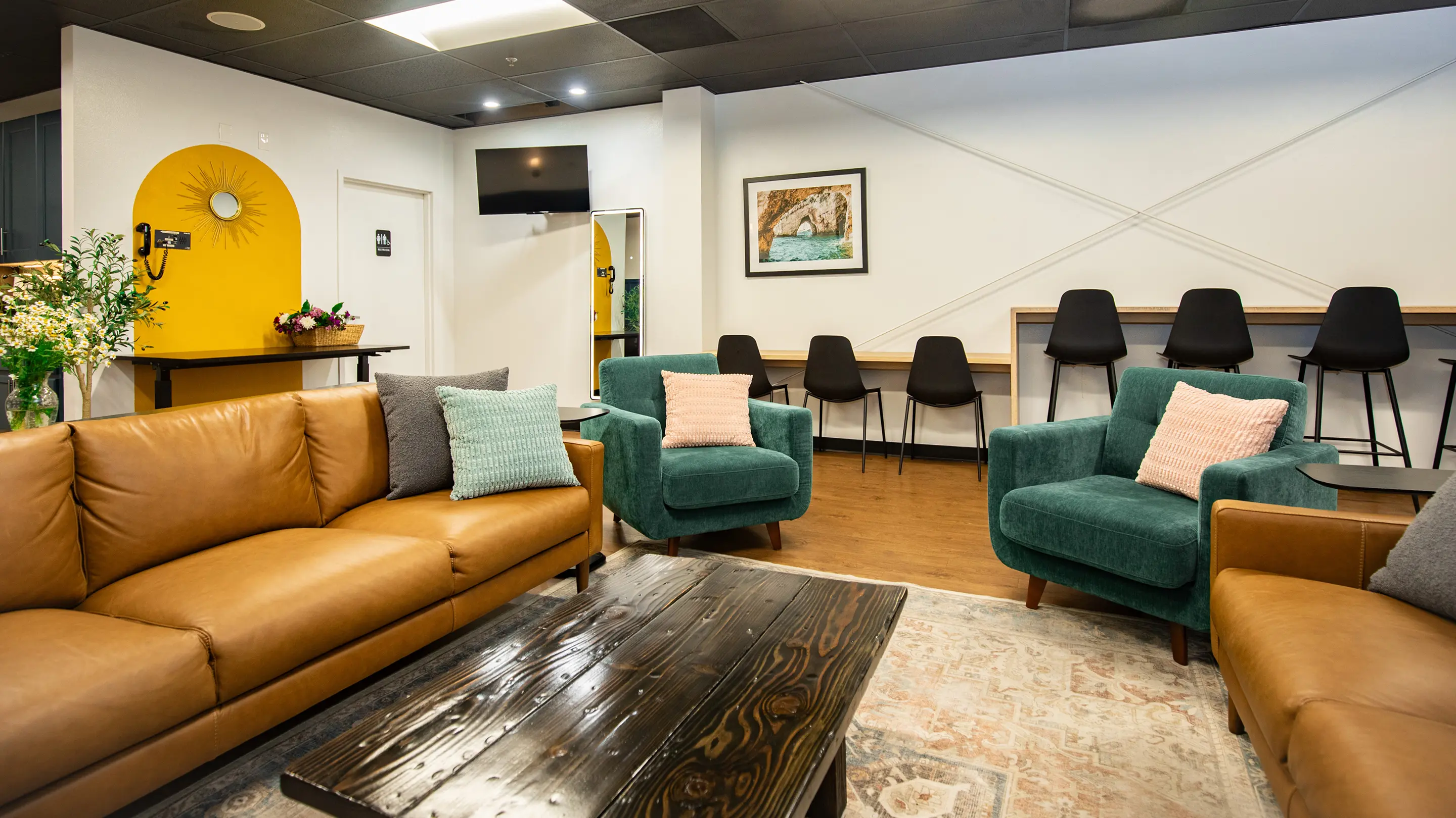 Eclectic lounge by Sunshine Collective Studio featuring cognac leather sofas, teal velvet armchairs, a reclaimed wood coffee table, vintage rug, oak bar counter, and a bold yellow arched alcove with sunburst mirror.