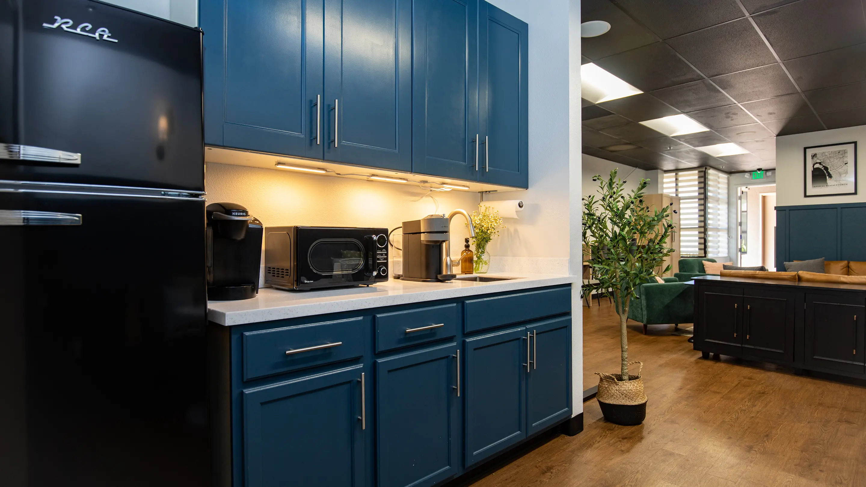 Commercial kitchenette by Sunshine Collective Studio with teal shaker cabinets, under-cabinet lighting, black retro refrigerator, and a view into the lounge with green velvet chairs and cognac leather sofa.