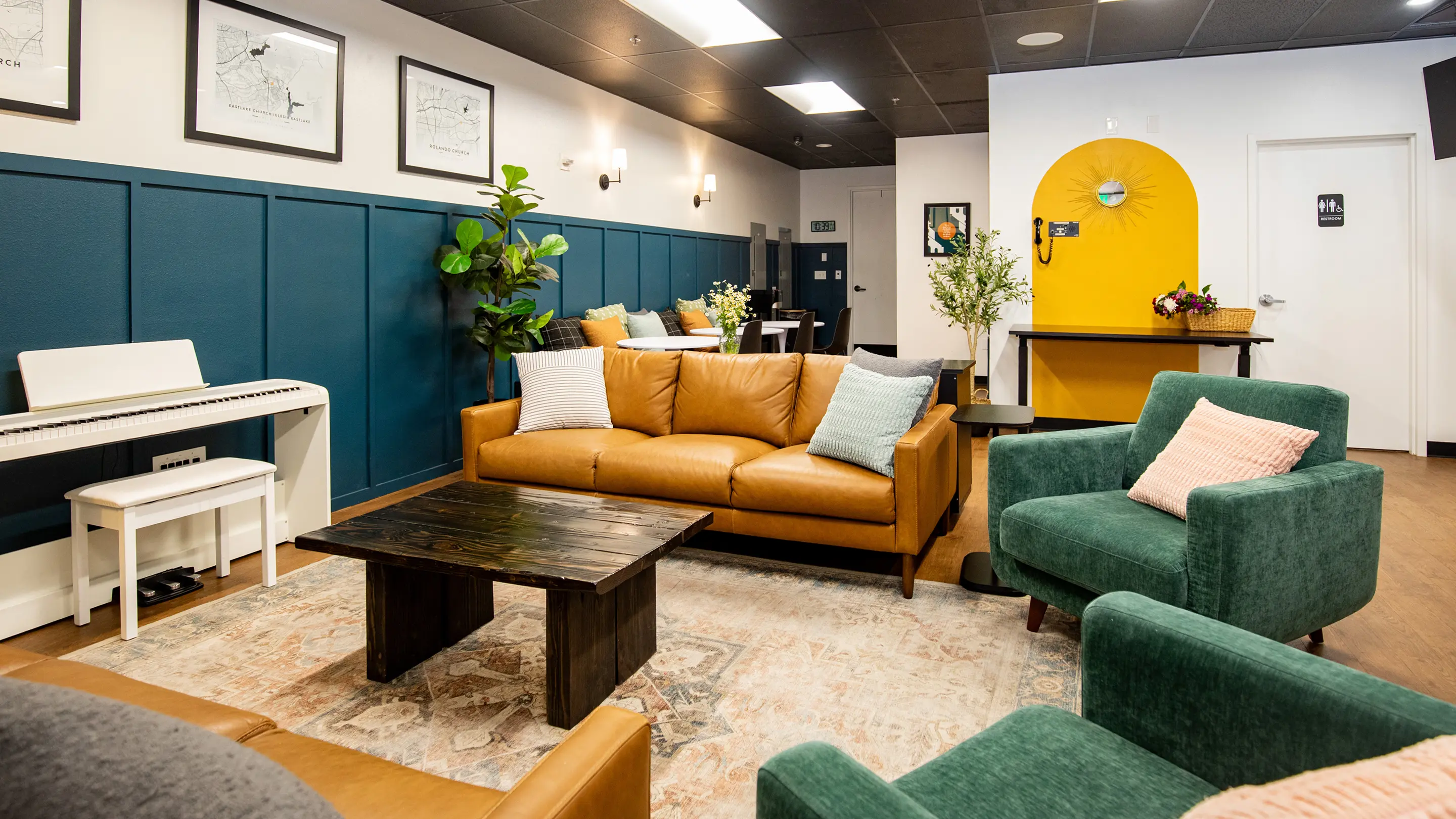 Eclectic lounge by Sunshine Collective Studio with a cognac leather sofa, green velvet chairs, reclaimed wood coffee table, teal board-and-batten walls, and a bold yellow arched alcove with sunburst mirror.