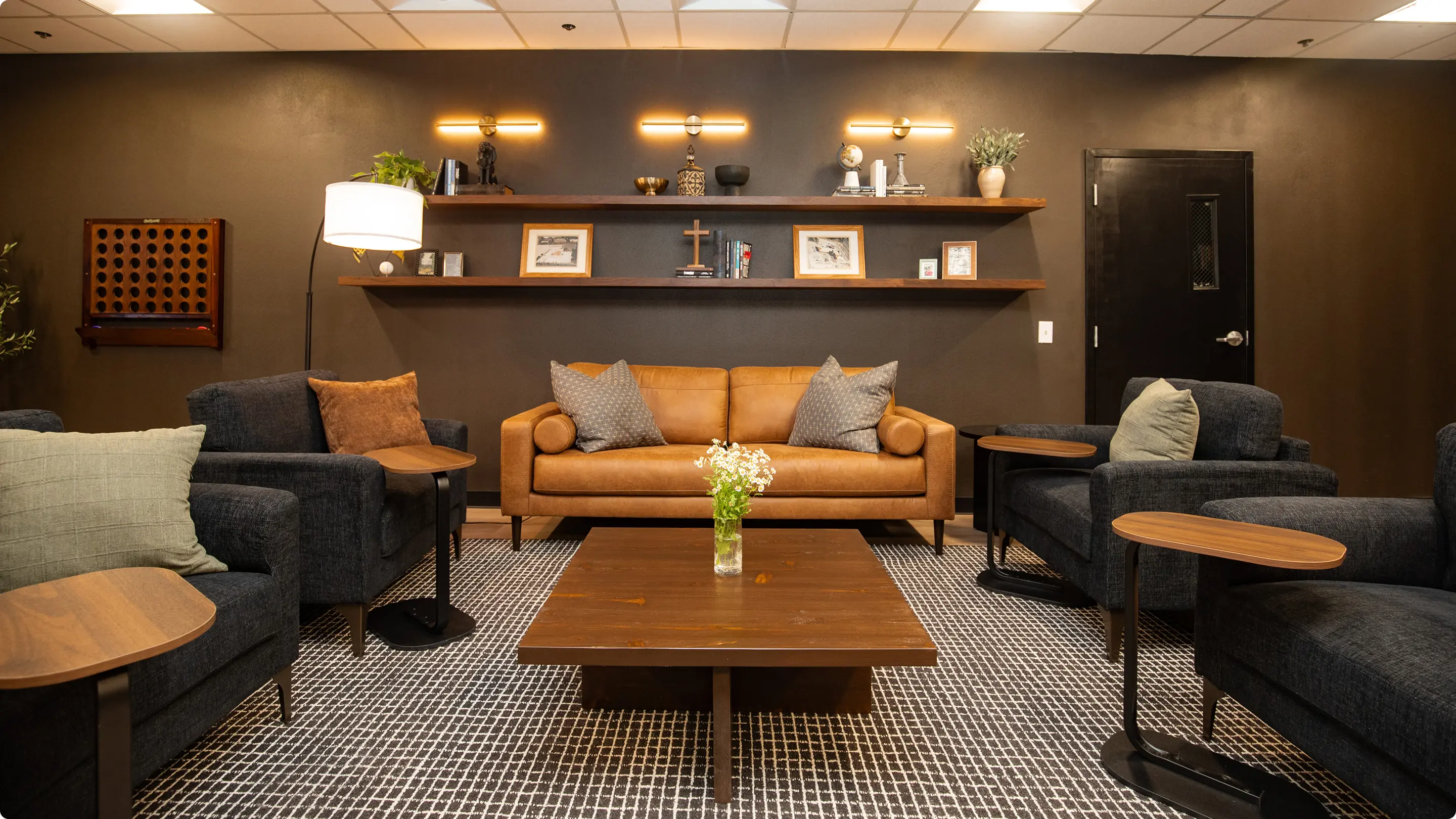 Commercial lounge by Sunshine Collective Studio with a cognac leather sofa, charcoal armchairs, walnut coffee table, and floating shelves with brass sconces on a deep charcoal accent wall. Grid-pattern rug and wall-mounted Connect Four add warmth.