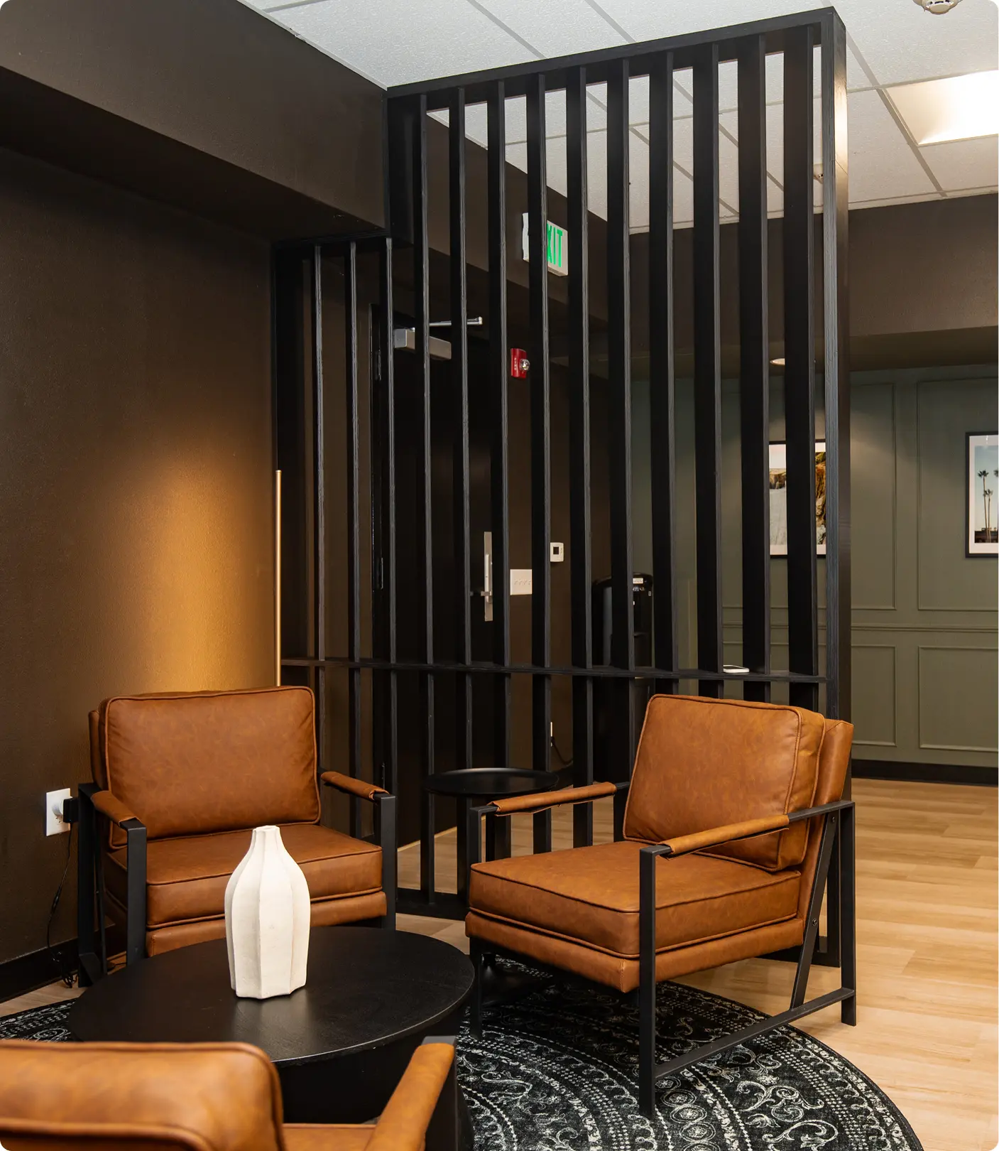 Lounge seating area by Sunshine Collective Studio with cognac leather armchairs and a dark drum table on a medallion rug, framed by a tall black vertical slat room divider against deep charcoal walls.