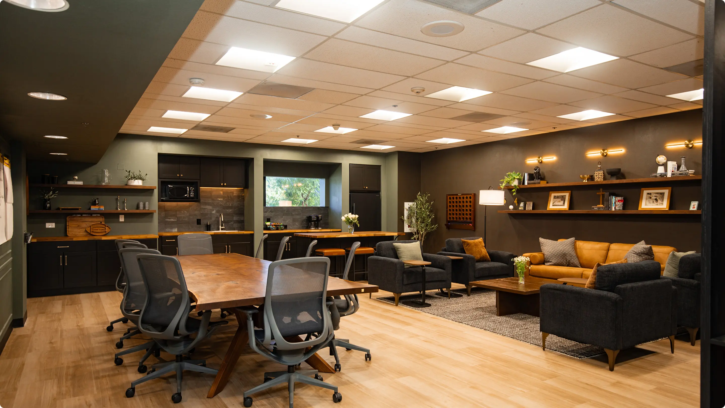 Open commercial space by Sunshine Collective Studio with a walnut conference table, mesh chairs, dark kitchenette with butcher block counters, and a lounge area with cognac leather sofa and charcoal armchairs against a charcoal accent wall.
