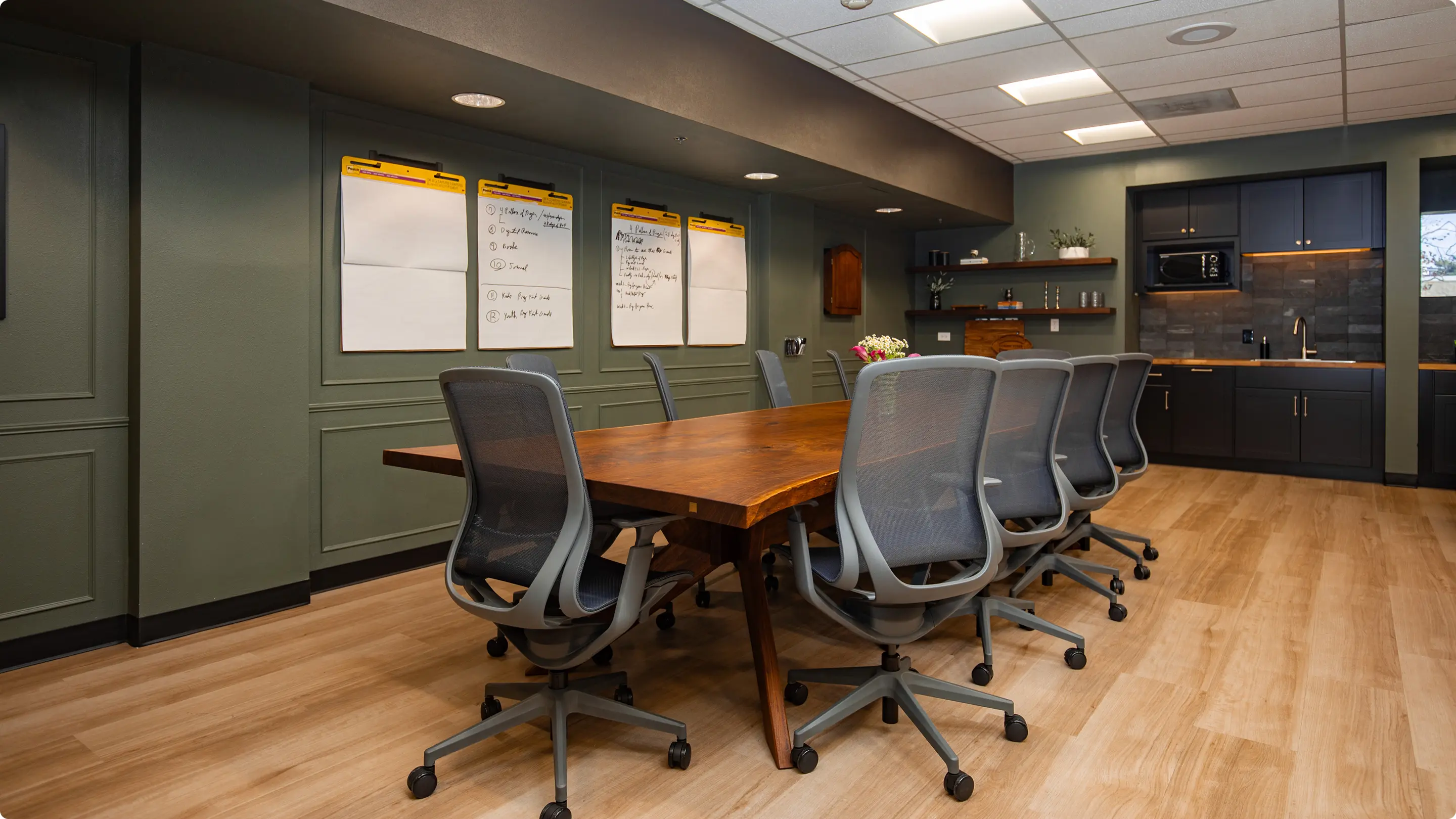 Conference room by Sunshine Collective Studio featuring a live-edge walnut table, mesh task chairs, sage green board-and-batten walls with flipchart paper, and a dark kitchenette with butcher block counters.