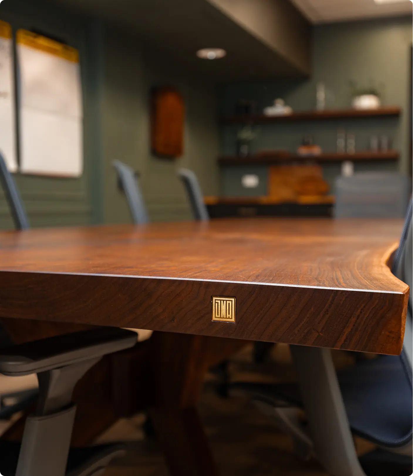 Close-up of a custom walnut conference table by Sunshine Collective Studio, featuring a brass maker's mark inset into the edge, with sage green wainscoting and wood shelving visible in the background.