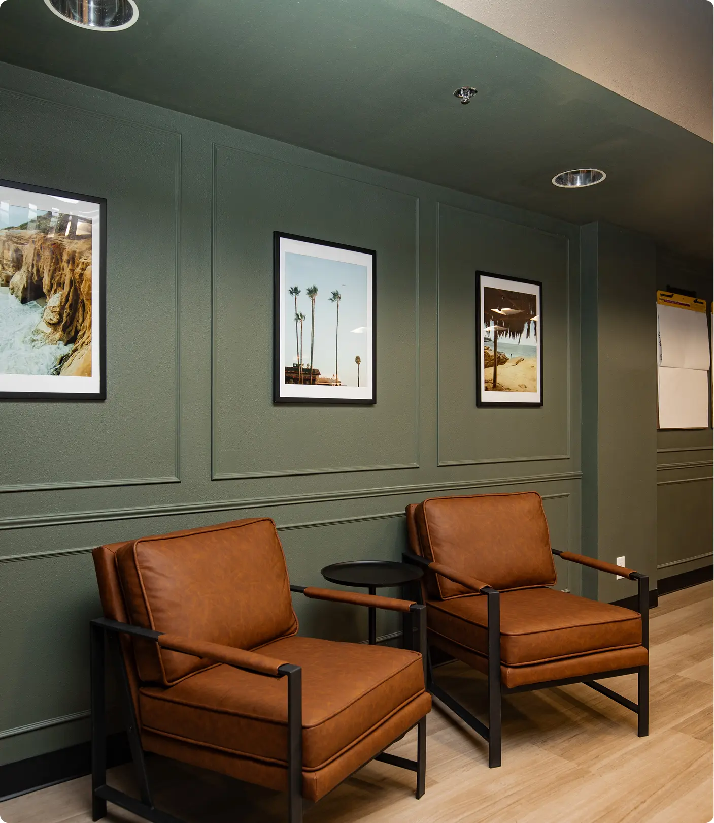 Hallway seating by Sunshine Collective Studio with two cognac leather armchairs and a black side table against sage green board-and-batten walls, accented by three framed coastal photography prints.