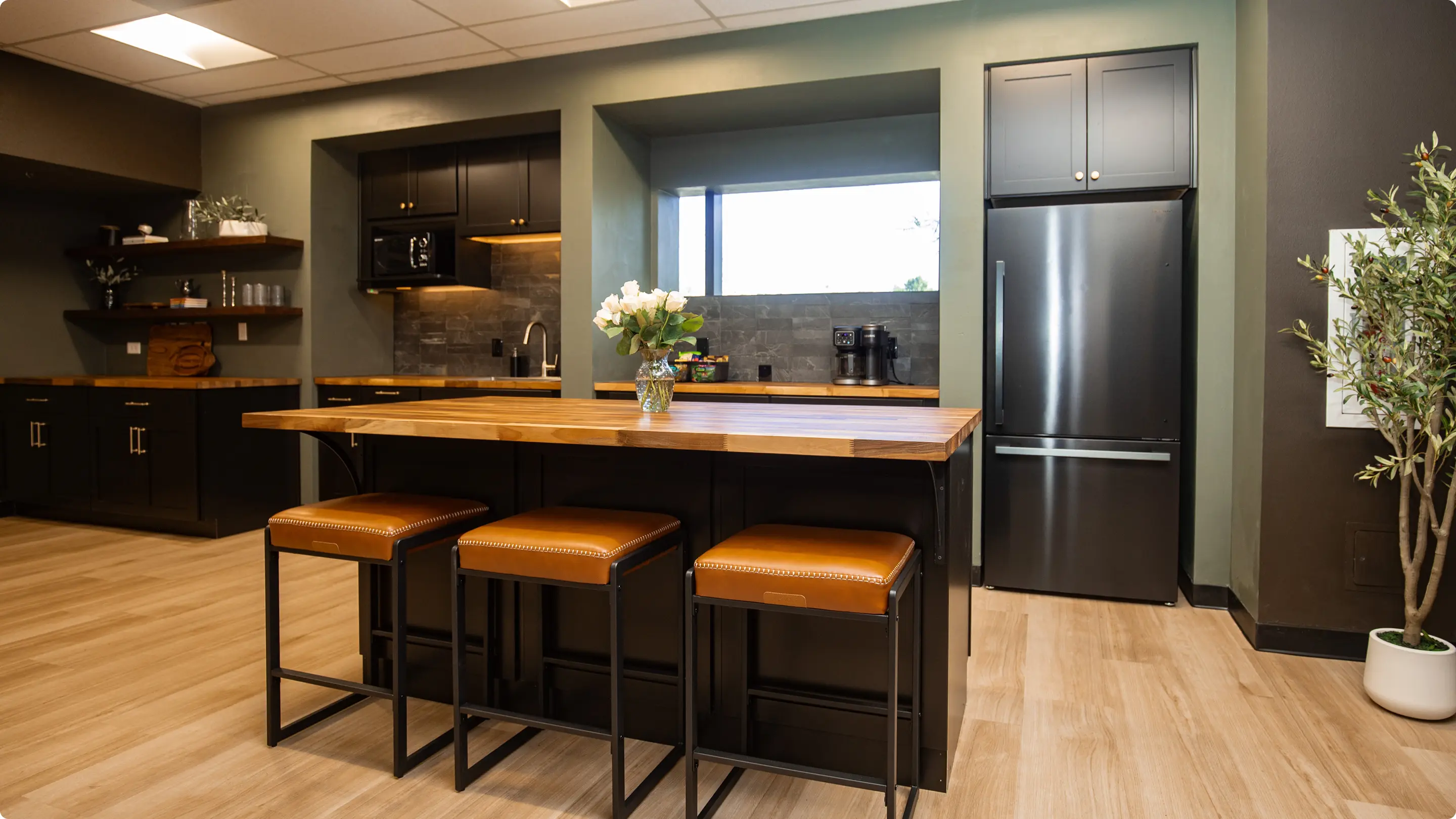 Commercial kitchen by Sunshine Collective Studio featuring dark shaker cabinets, butcher block counters and island with cognac leather stools, slate tile backsplash, black refrigerator, and an olive tree on light hardwood floors.