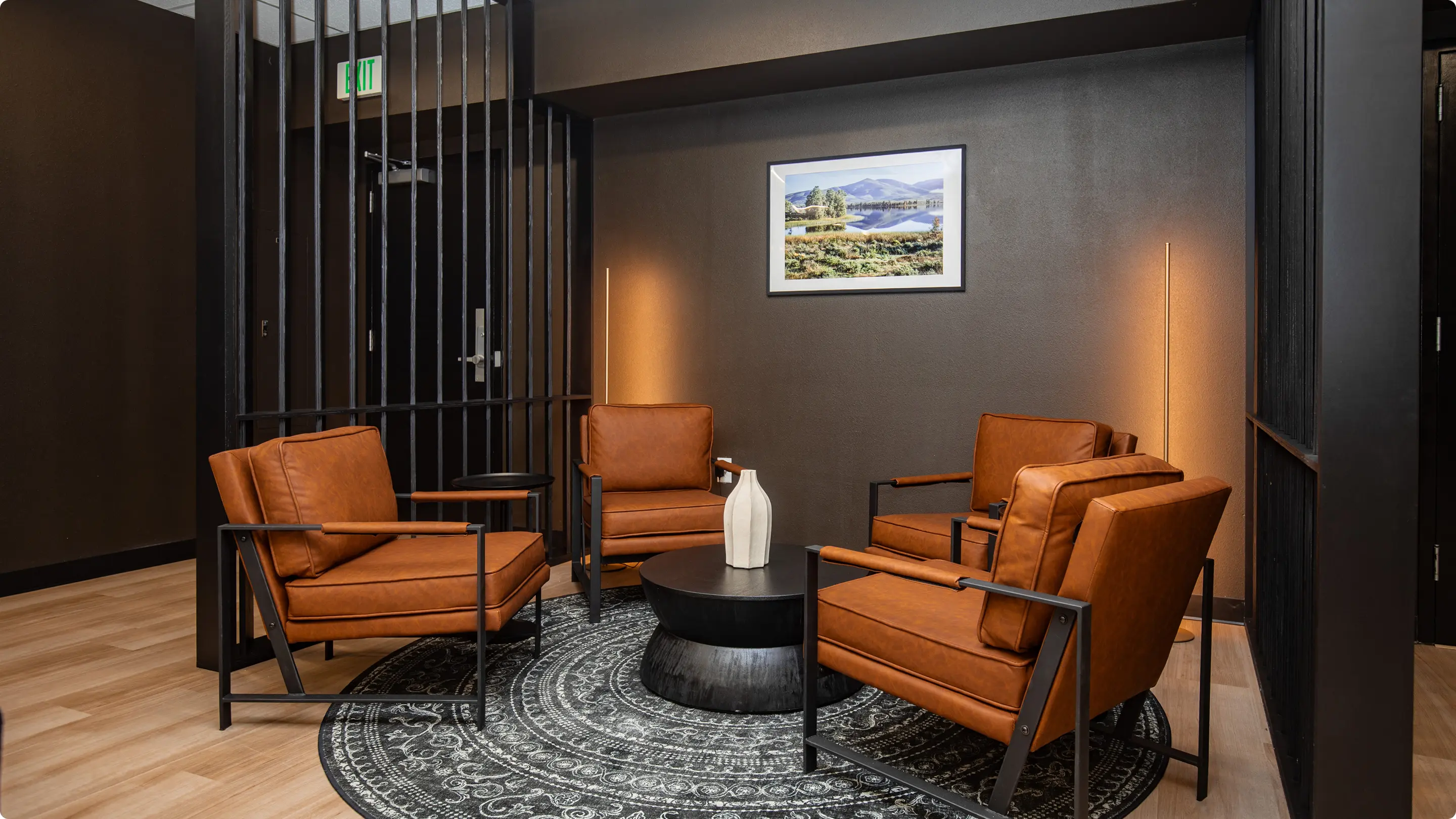 Private lounge nook by Sunshine Collective Studio with four cognac leather armchairs around a dark drum coffee table on a medallion rug, framed by black vertical slat dividers and a landscape print on charcoal walls.