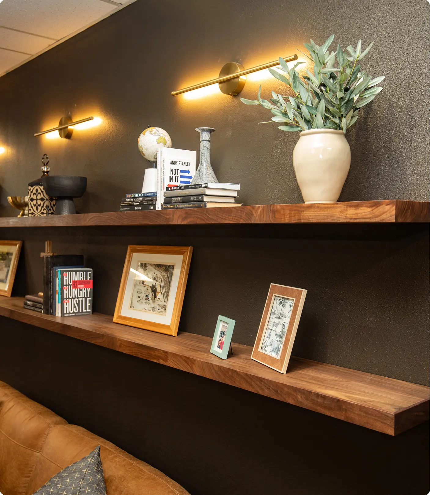 Floating walnut shelf detail by Sunshine Collective Studio, styled with books, a globe, ceramic vessels, framed photos, and an olive branch arrangement under warm brass bar sconces on a charcoal wall.