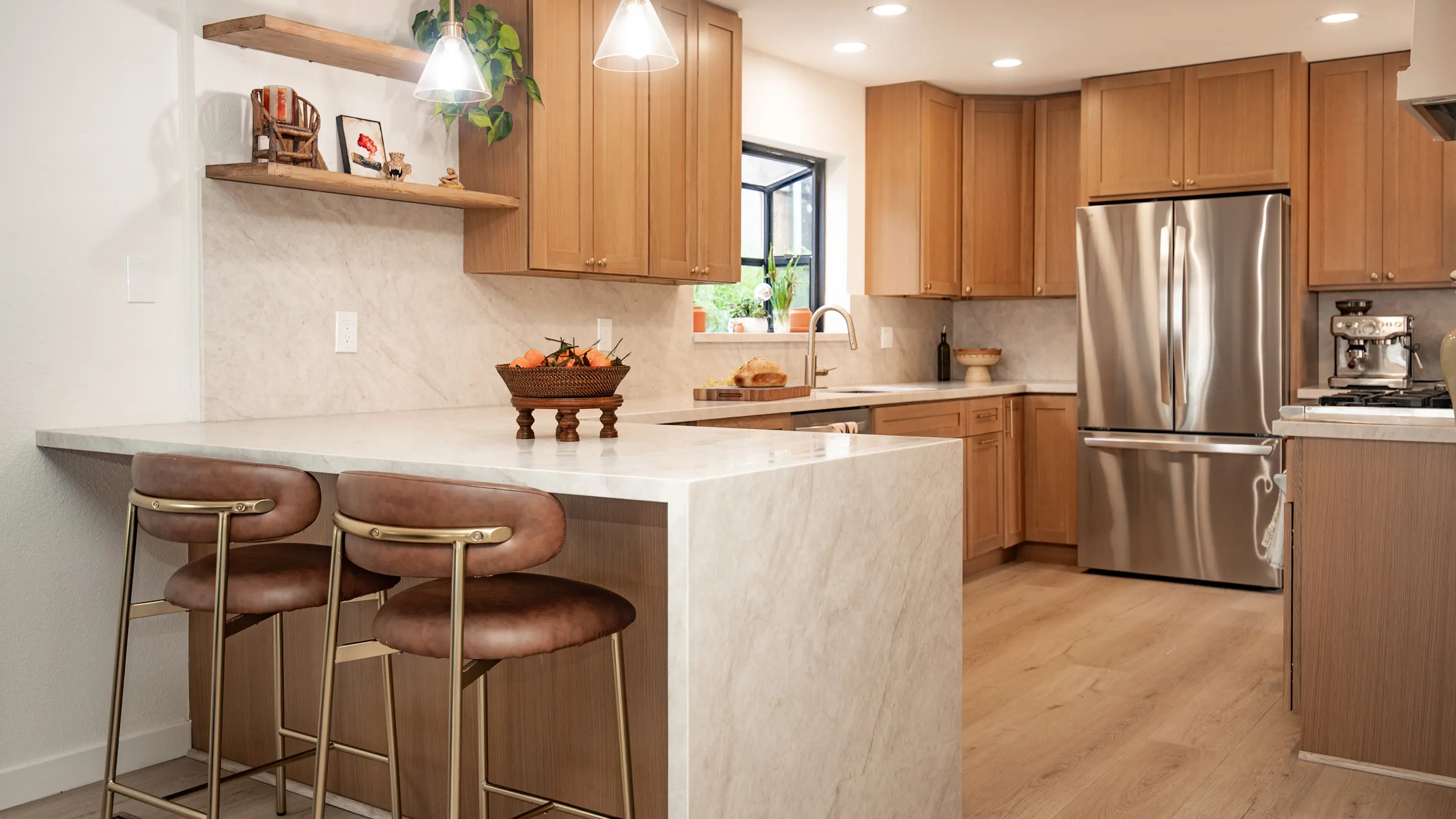 Renovated kitchen by Sunshine Collective Studio with warm honey shaker cabinets, marble waterfall island, leather-and-brass bar stools, glass pendant lights, floating wood shelves, and full marble slab backsplash on light hardwood floors.