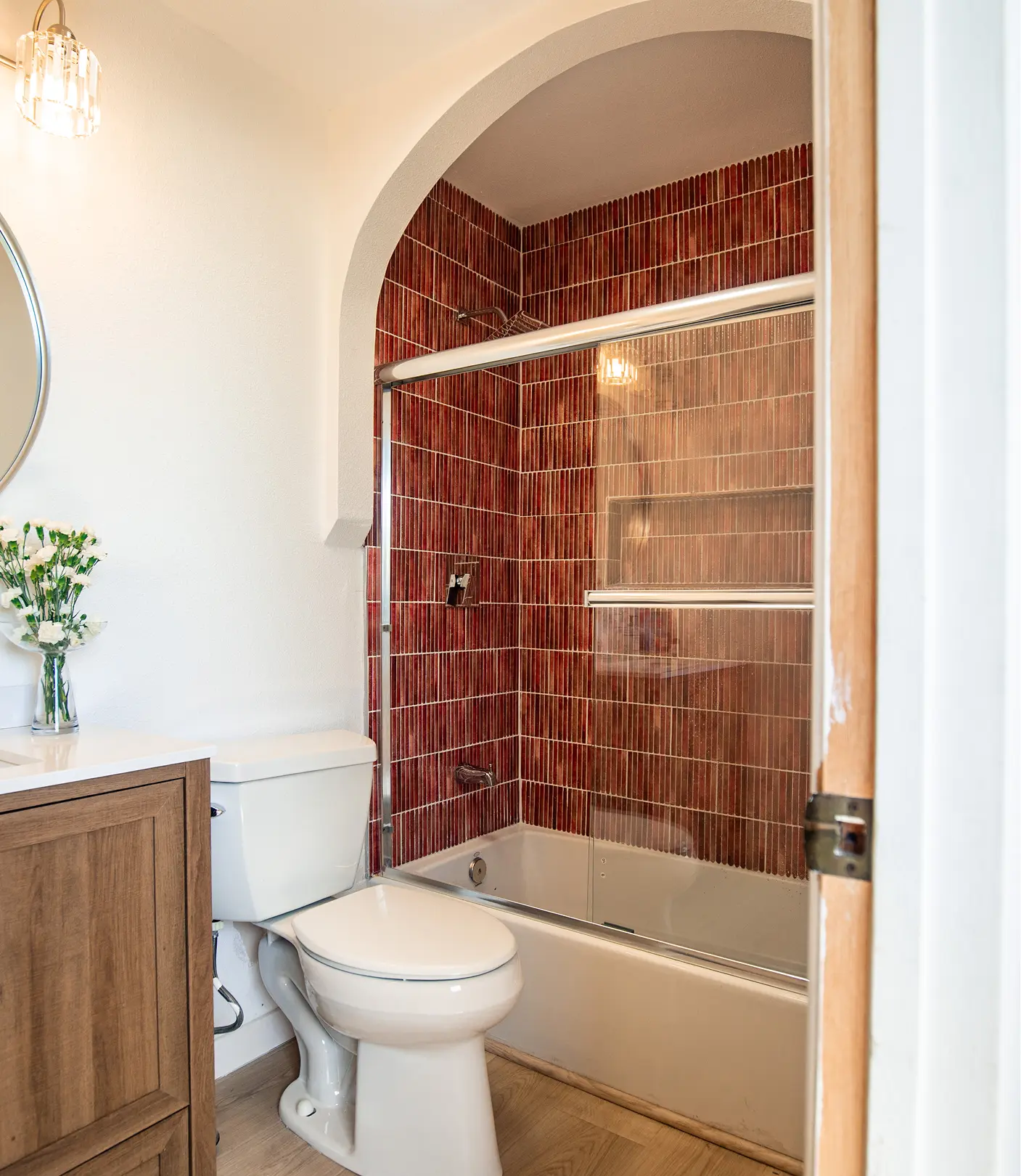 Bathroom by Sunshine Collective Studio featuring a bold arched tub-shower alcove tiled in deep terracotta finger tile, wood-tone vanity, crystal sconce, and a round mirror on white walls.