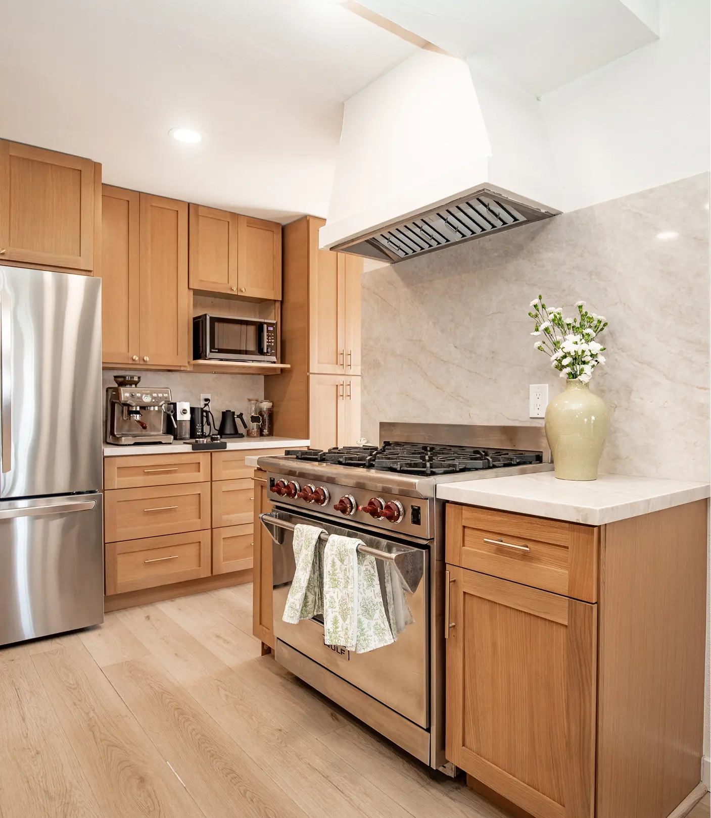 Kitchen by Sunshine Collective Studio featuring a professional stainless range with hood, warm shaker cabinets, marble backsplash, built-in microwave, and a full coffee and espresso station on light hardwood floors.