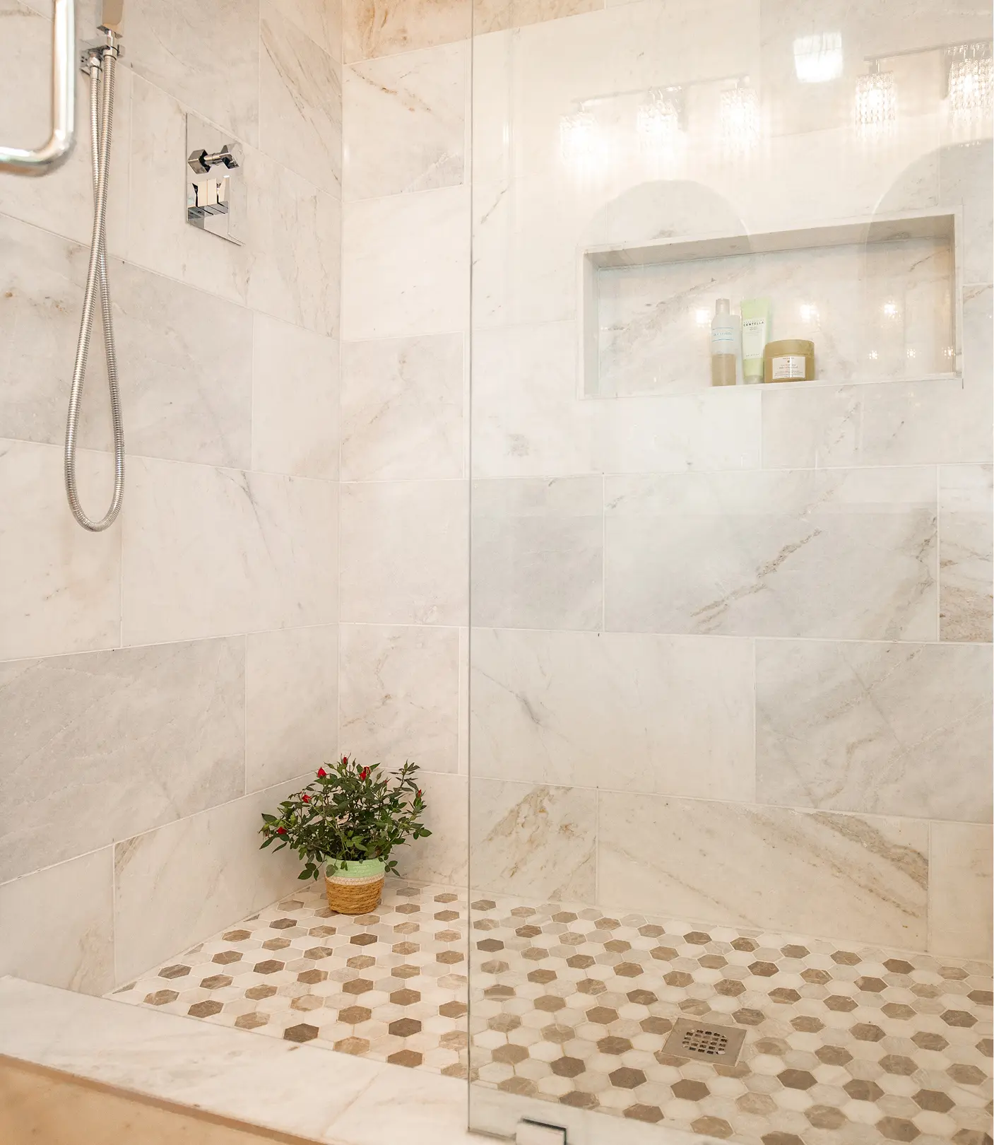 Walk-in shower by Sunshine Collective Studio with floor-to-ceiling marble tile, hexagon mosaic floor, built-in niche, chrome handheld showerhead, and a small potted plant.