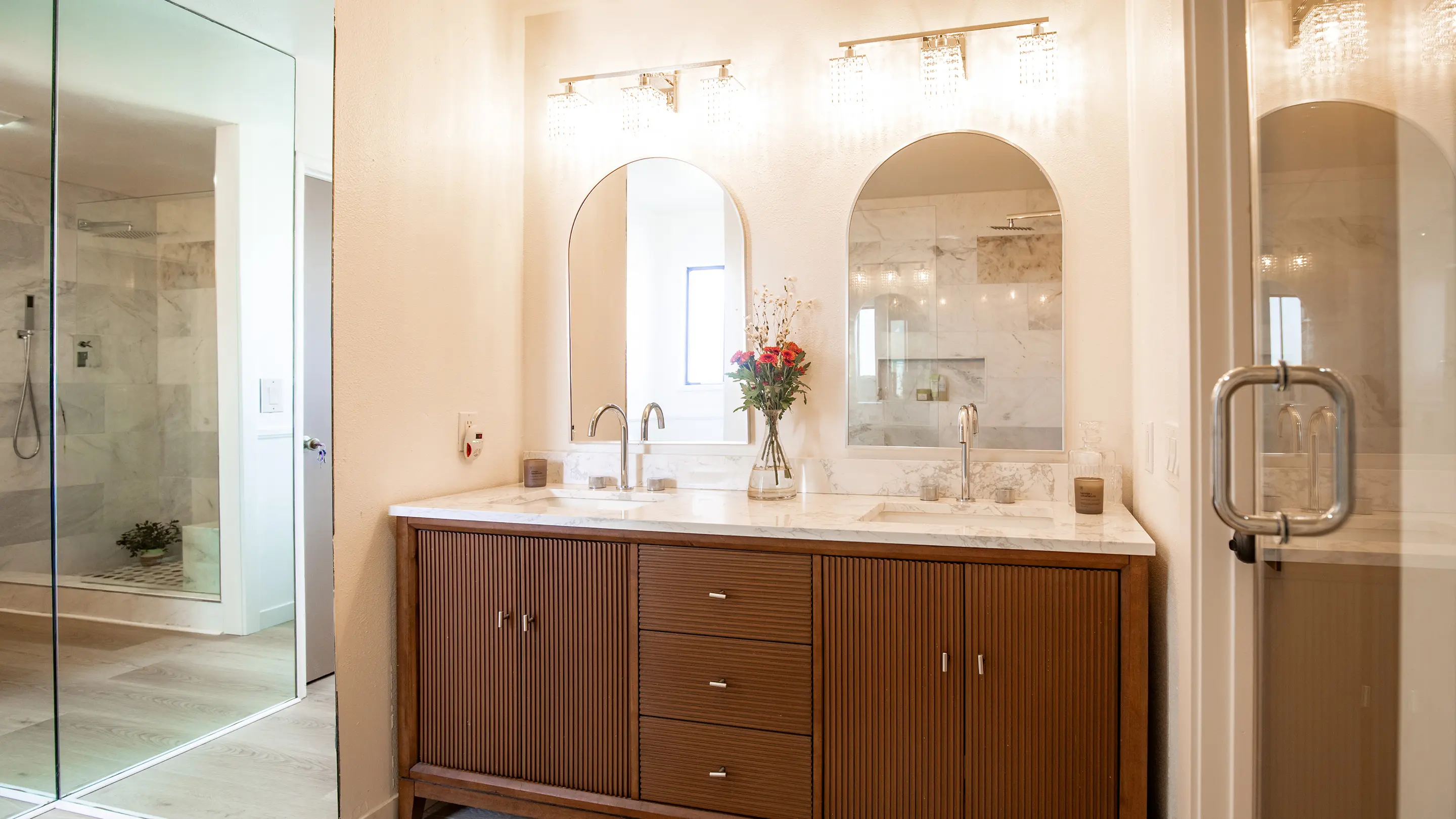 Primary bathroom by Sunshine Collective Studio featuring dual arched mirrors with crystal sconces, fluted walnut double vanity, marble countertop, and a glass-enclosed marble shower with rain head.